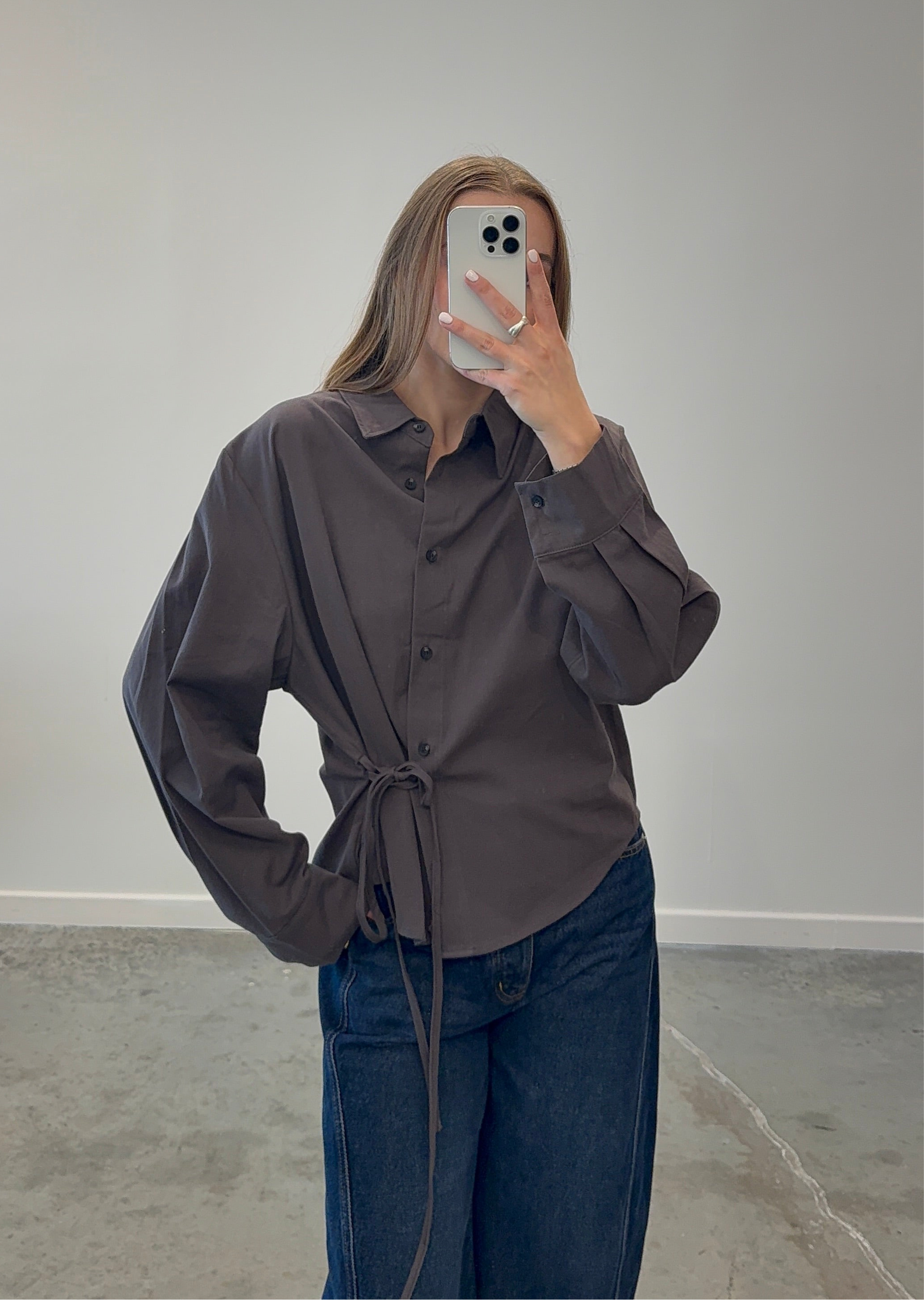 Woman wearing a dark brown shirt with side tie and blue jeans taking a mirror selfie. Fall fashion