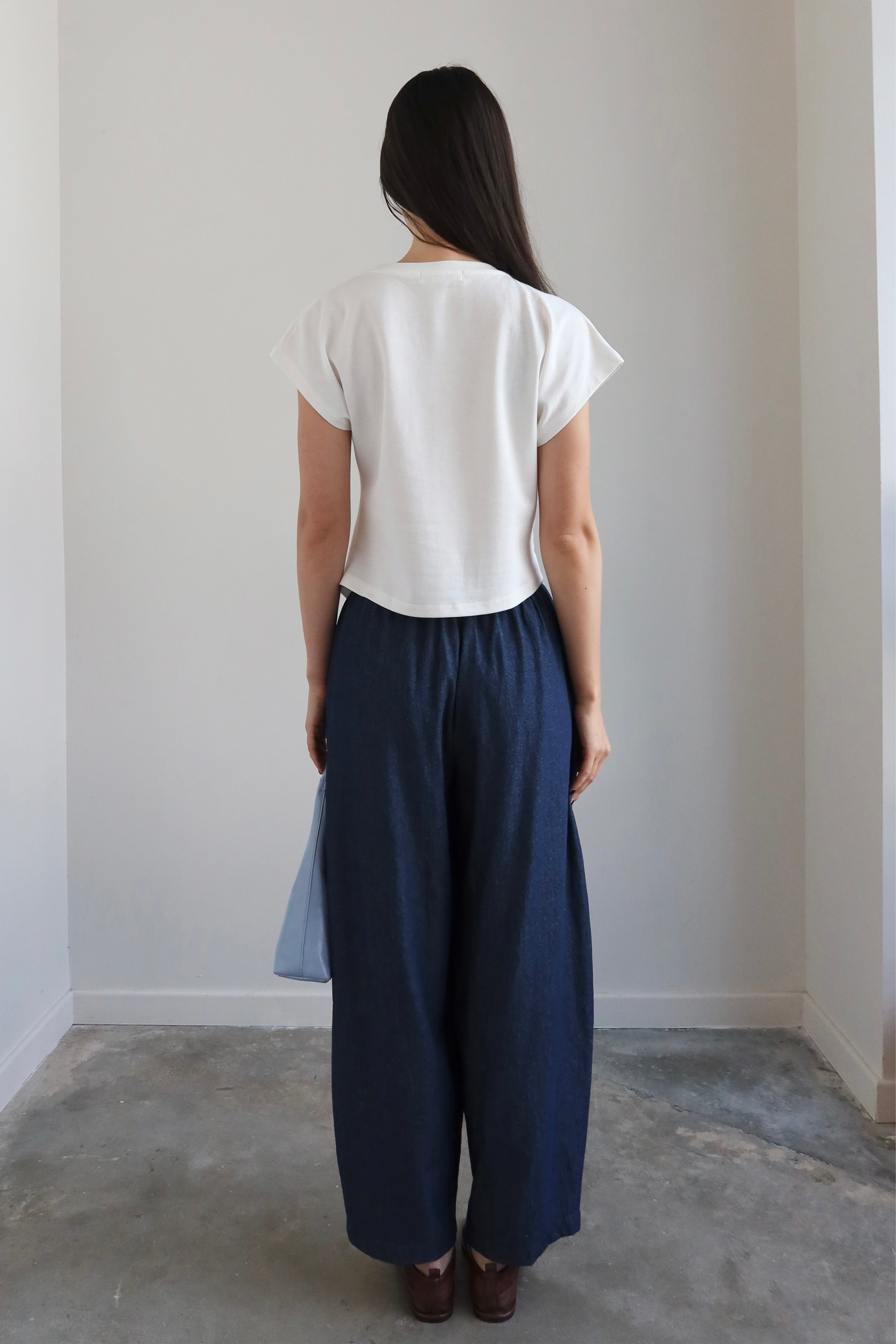 Woman wearing a white top and blue pants standing against a plain wall.