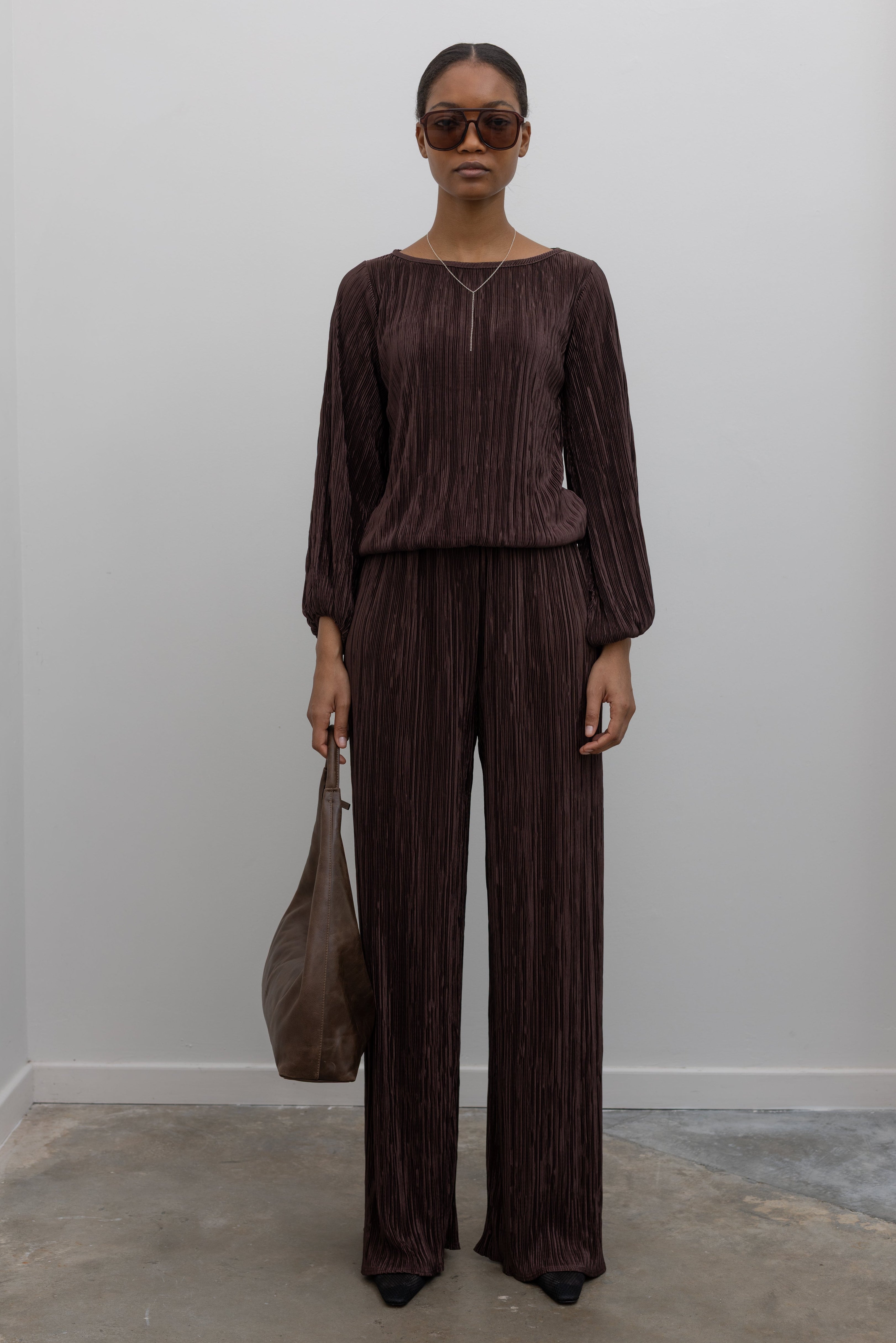 Beautiful woman wearing a brown plisse outfit with sunglasses holding a marge sherwood bag at alas eius