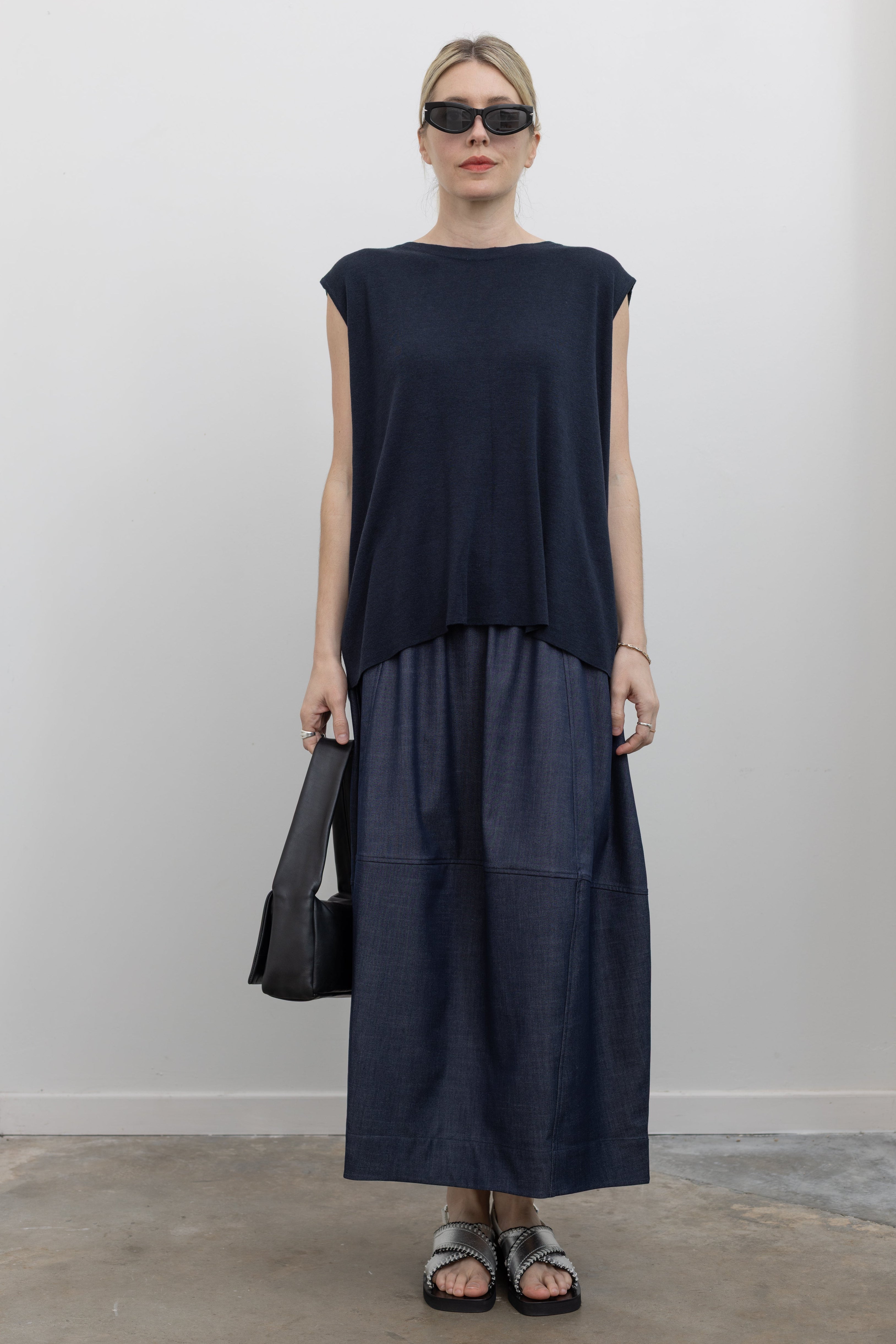 Full length front view of a woman standing wearing a modern and modest Mijeong Park brand navy blue sleeveless top with a fluid draped back and a curved front hem paired with a dark wash denim midi skirt at an Alas Eius store. 