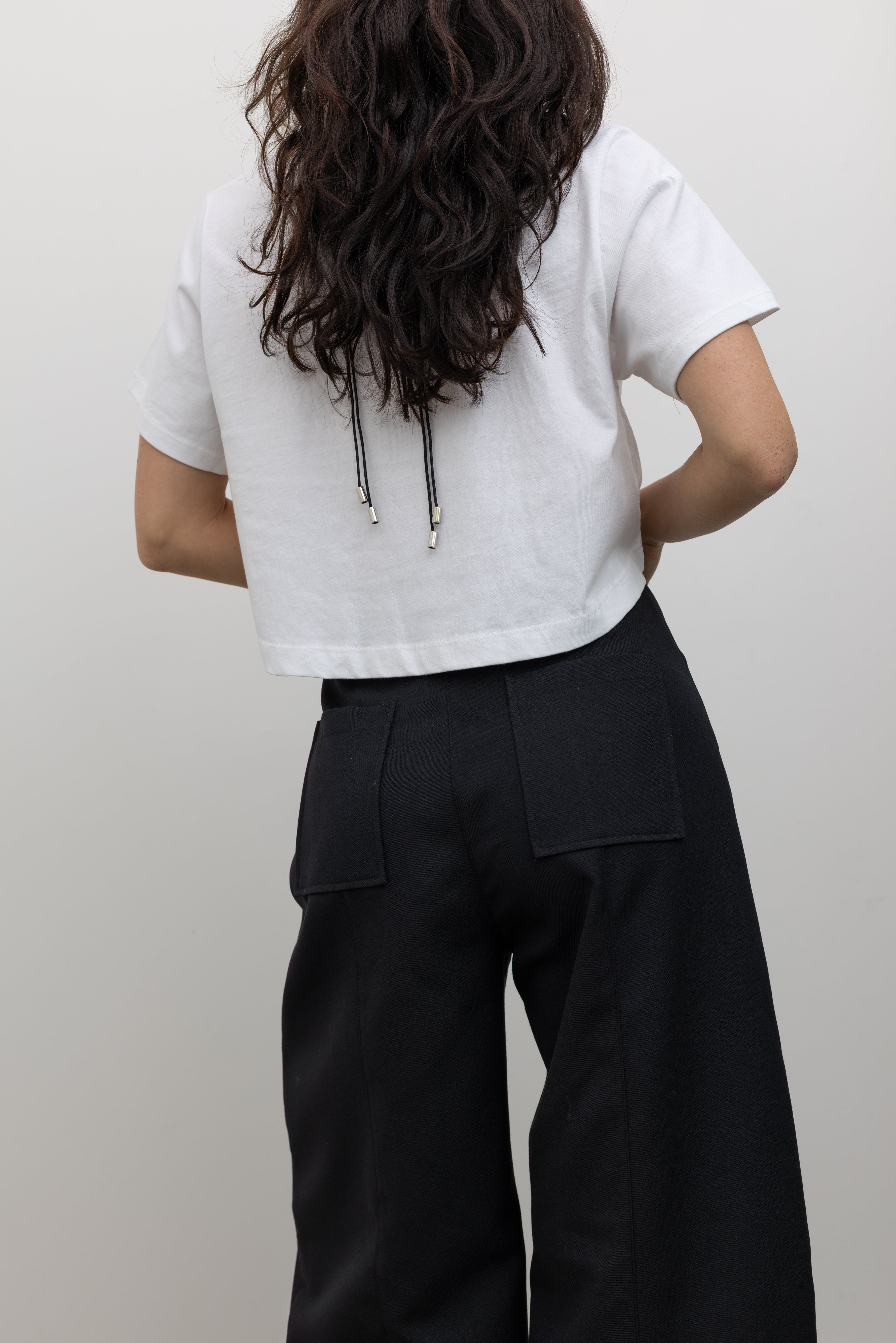 Close-up back view of a woman standing wearing the perfect versatile workwear pant in an Alas Eius Label black wool carrot silhouette high-waisted, wide-leg trouser paired with an Alas Eius label white cotton boxy cropped top at Alas Eius. 