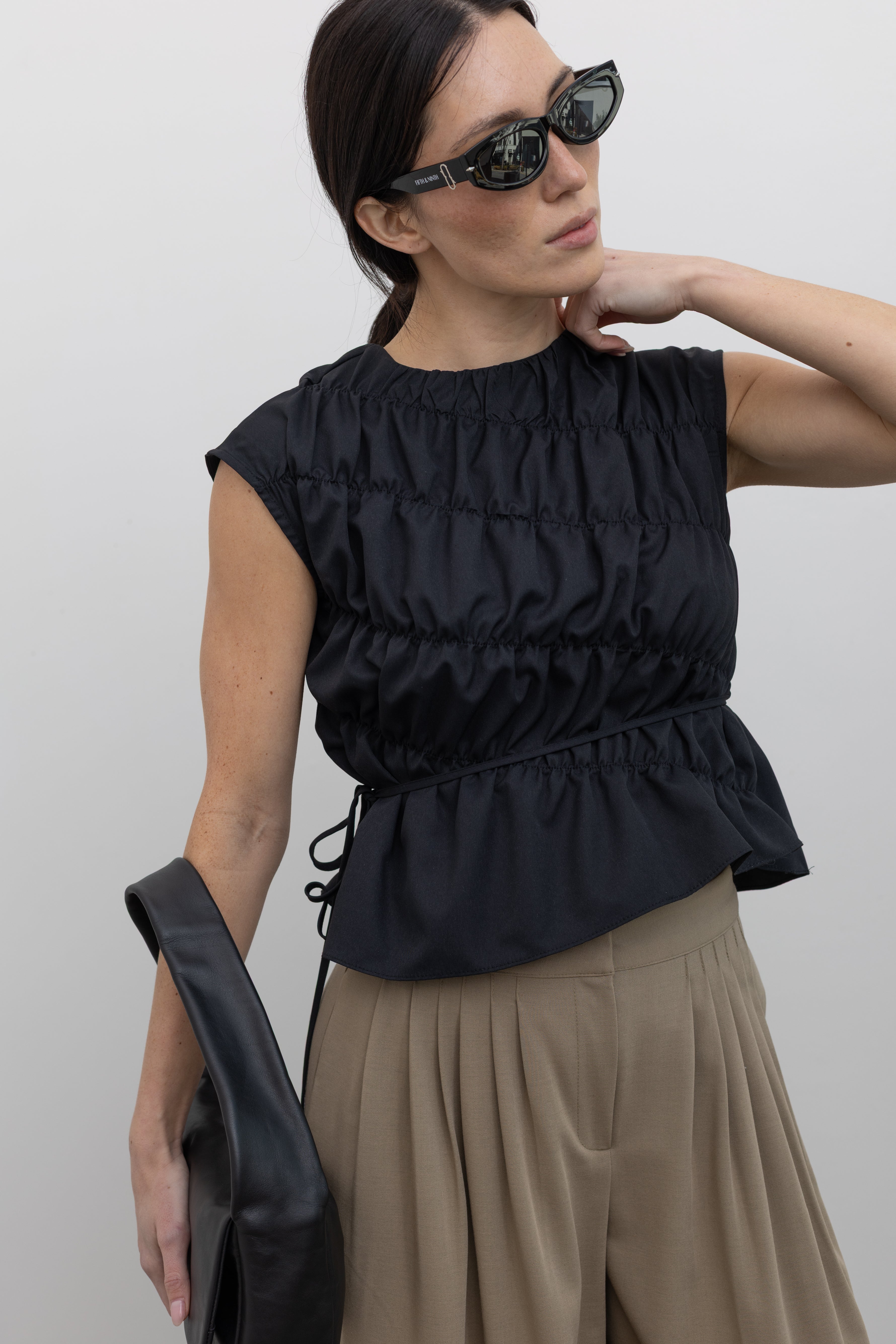 Front view of a woman posing wearing contemporary and chic womens fashion in an Alas Eius Black Asymmetrical Cinched Top featuring a crew neckline, cap sleeves, and an optional waist tie at an Alas Eius store. 