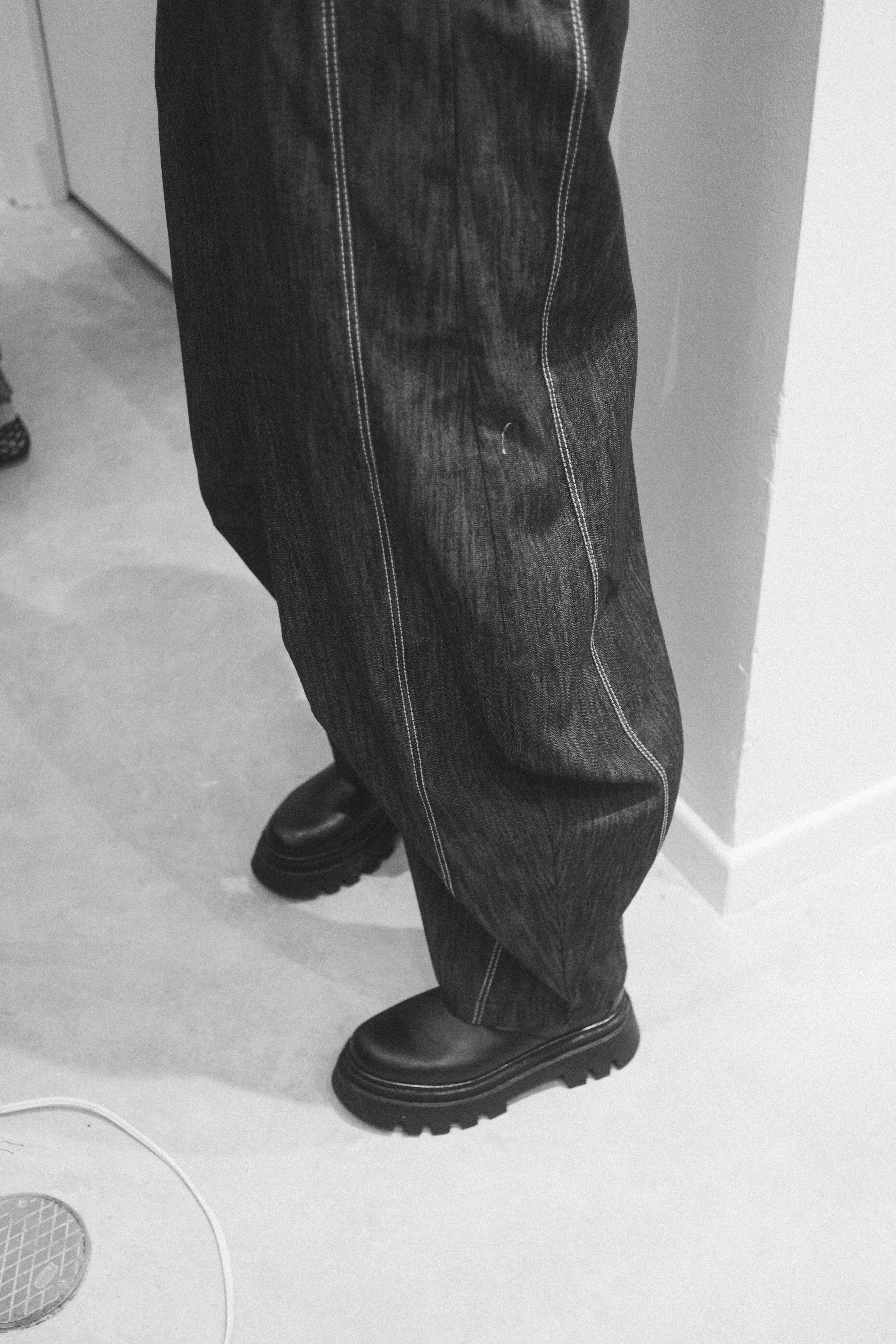 A person wearing black barrel-leg pants with contrast stitching and a pair of black shoes.