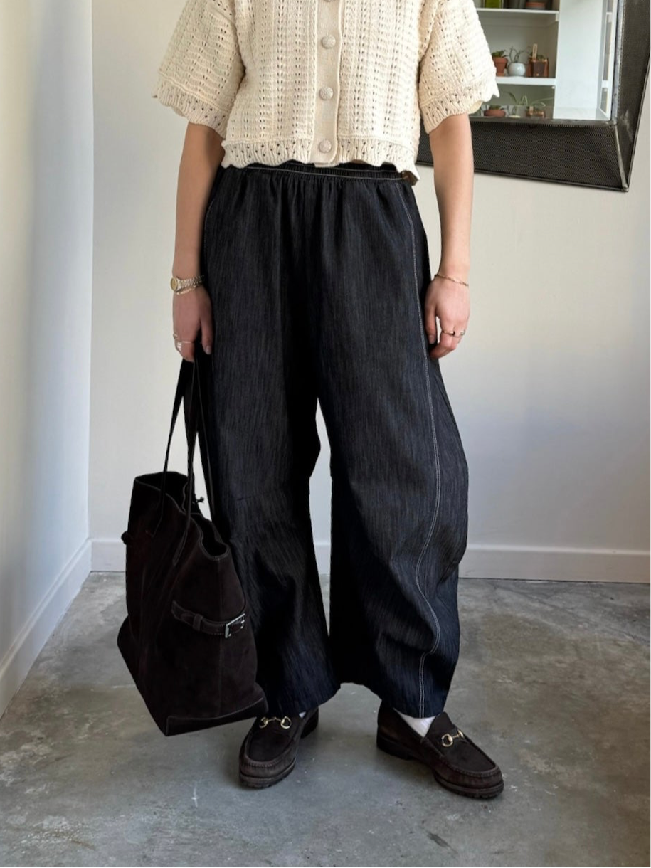 A person standing in an indoor setting wearing black barrel jeans with a relaxed fit and elastic waistband, featuring double contrast stitching, paired with a beige lace top and black shoes.