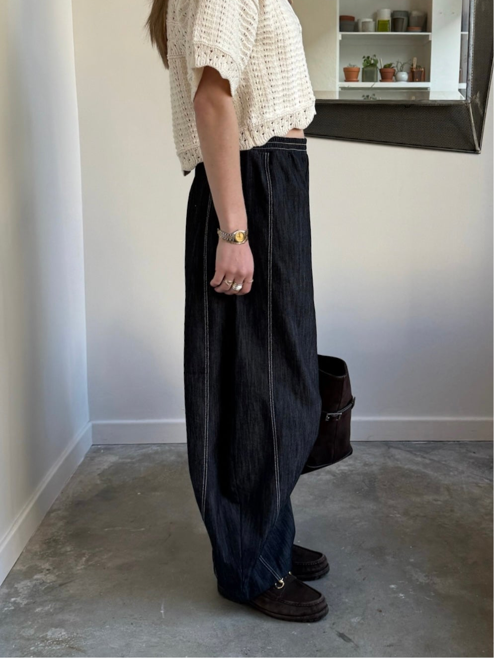 A person standing in an indoor setting wearing black barrel jeans with a relaxed fit and elastic waistband, featuring double contrast stitching, paired with a beige lace top and black shoes.