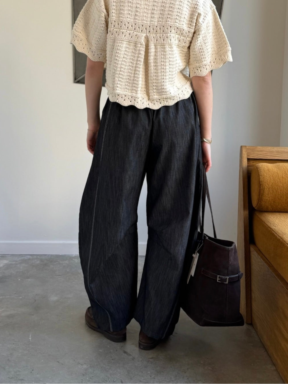 A person standing in an indoor setting wearing black barrel jeans with a relaxed fit and elastic waistband, featuring double contrast stitching, paired with a beige lace top and black shoes.