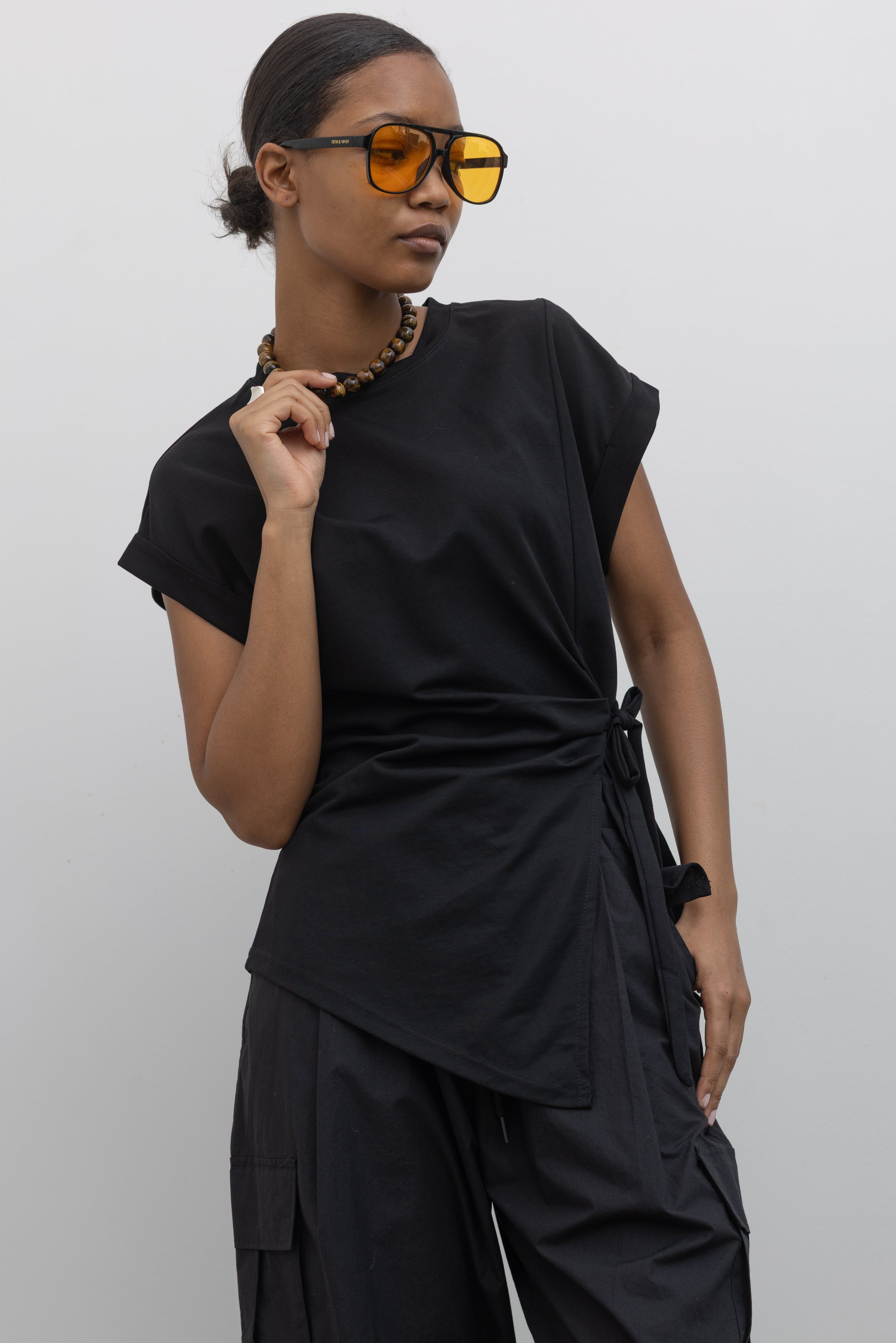 Woman wearing essential capsule wardrobe staple in an Alas Eius Black Drop Shoulder Ribbon Side Tie Cotton Top with black cargo pants and sunglasses at Alas Eius