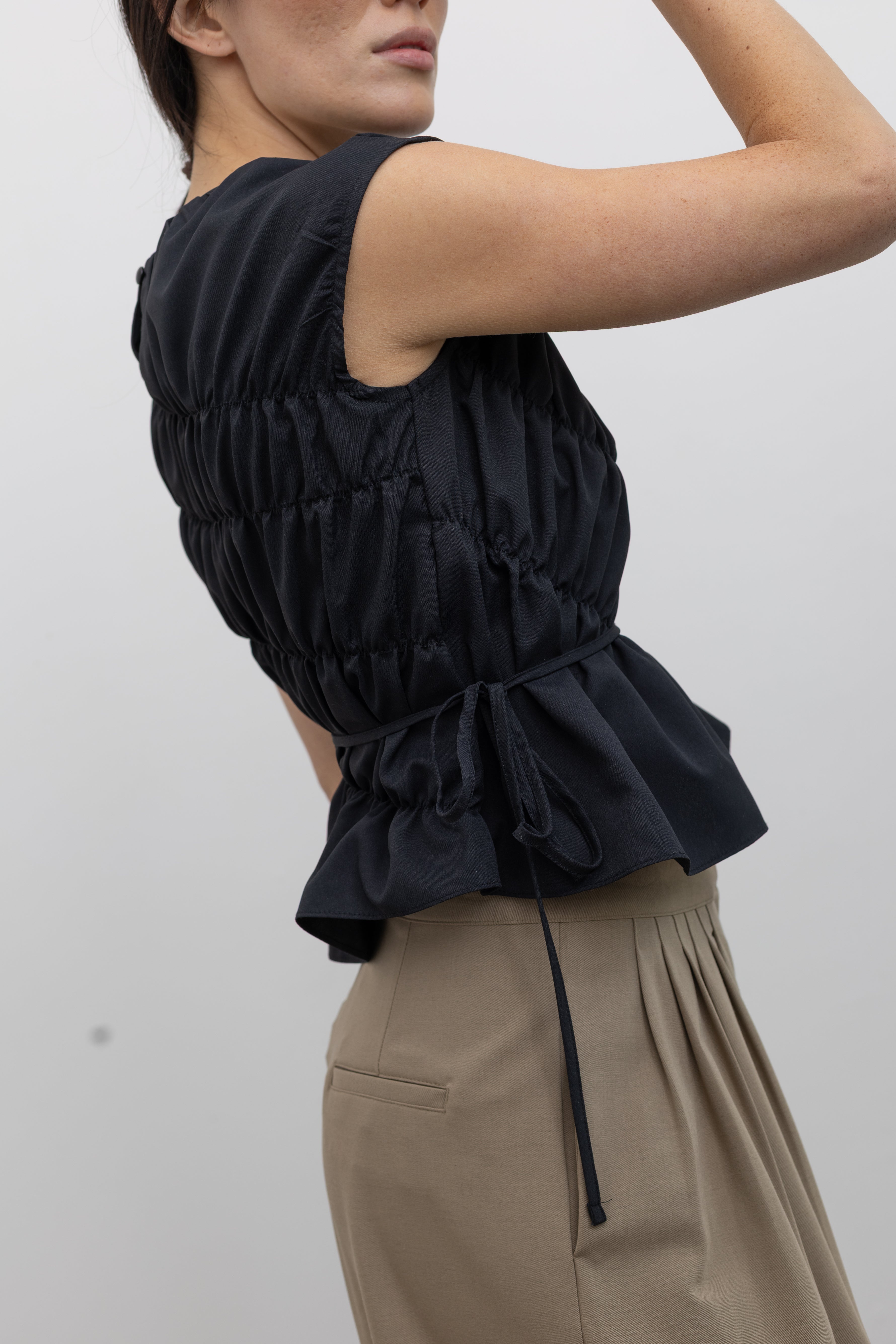 Close-up side view of a woman posing wearing contemporary and chic women's fashion in an Alas Eius Black Asymmetrical Cinched Top featuring a crew neckline, cap sleeves, and an optional waist tie at an Alas Eius store. 