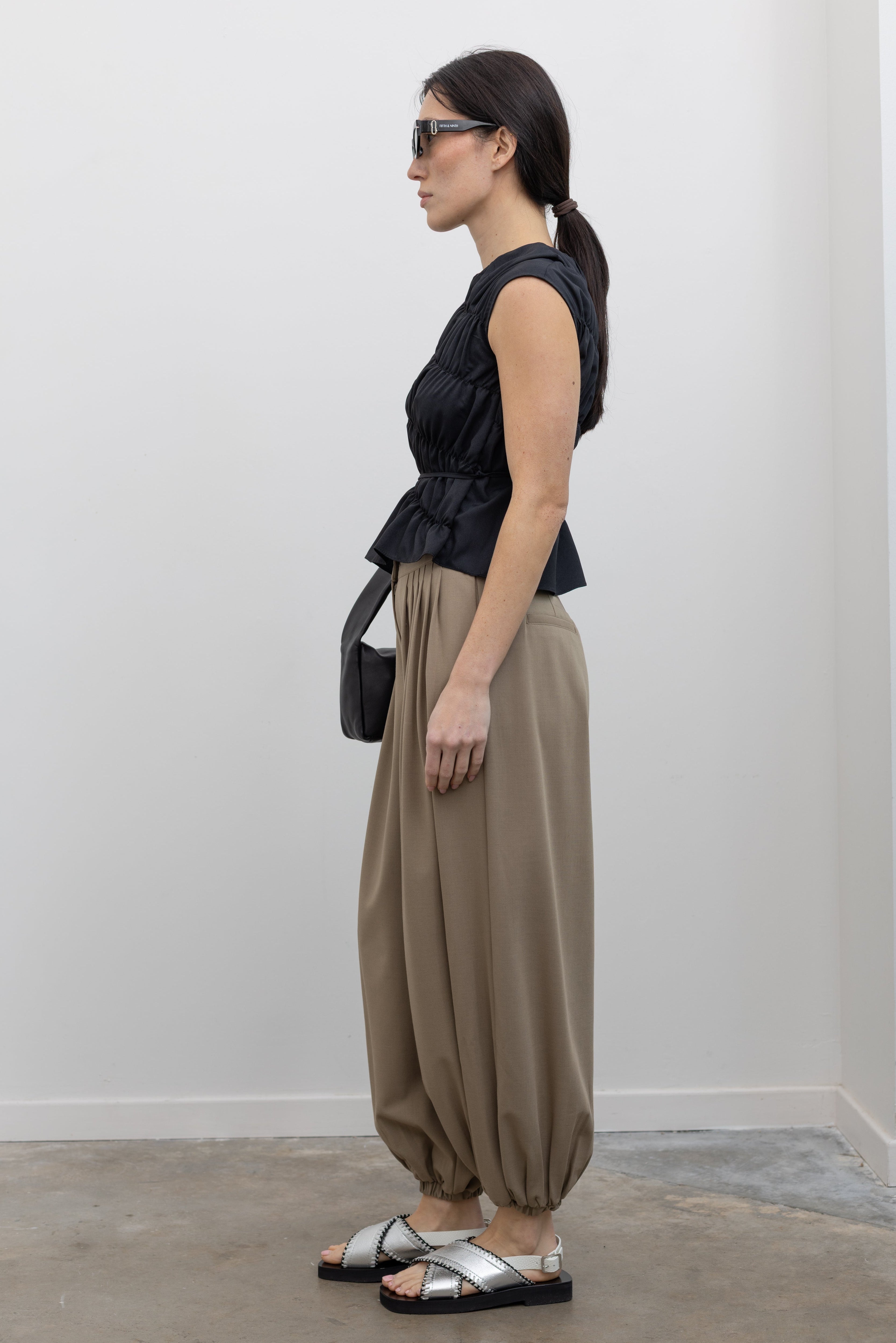 Side view of a woman posing wearing contemporary and chic women's fashion in an Alas Eius Black Asymmetrical Cinched Top featuring a crew neckline, cap sleeves, and an optional waist tie paired with elevated khaki cinched ankle pants, silver sandals, and a black leather St. Agni bag at an Alas Eius store. 