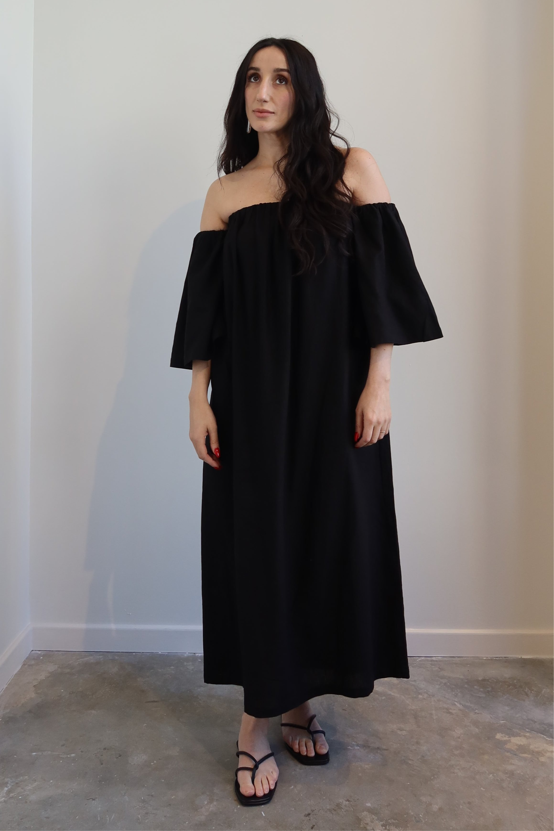 A woman standing, wearing an off-the-shoulder black midi dress with a flowy silhouette, sleeveless design, and side slant pockets. She has dark hair and is posing against a neutral background.