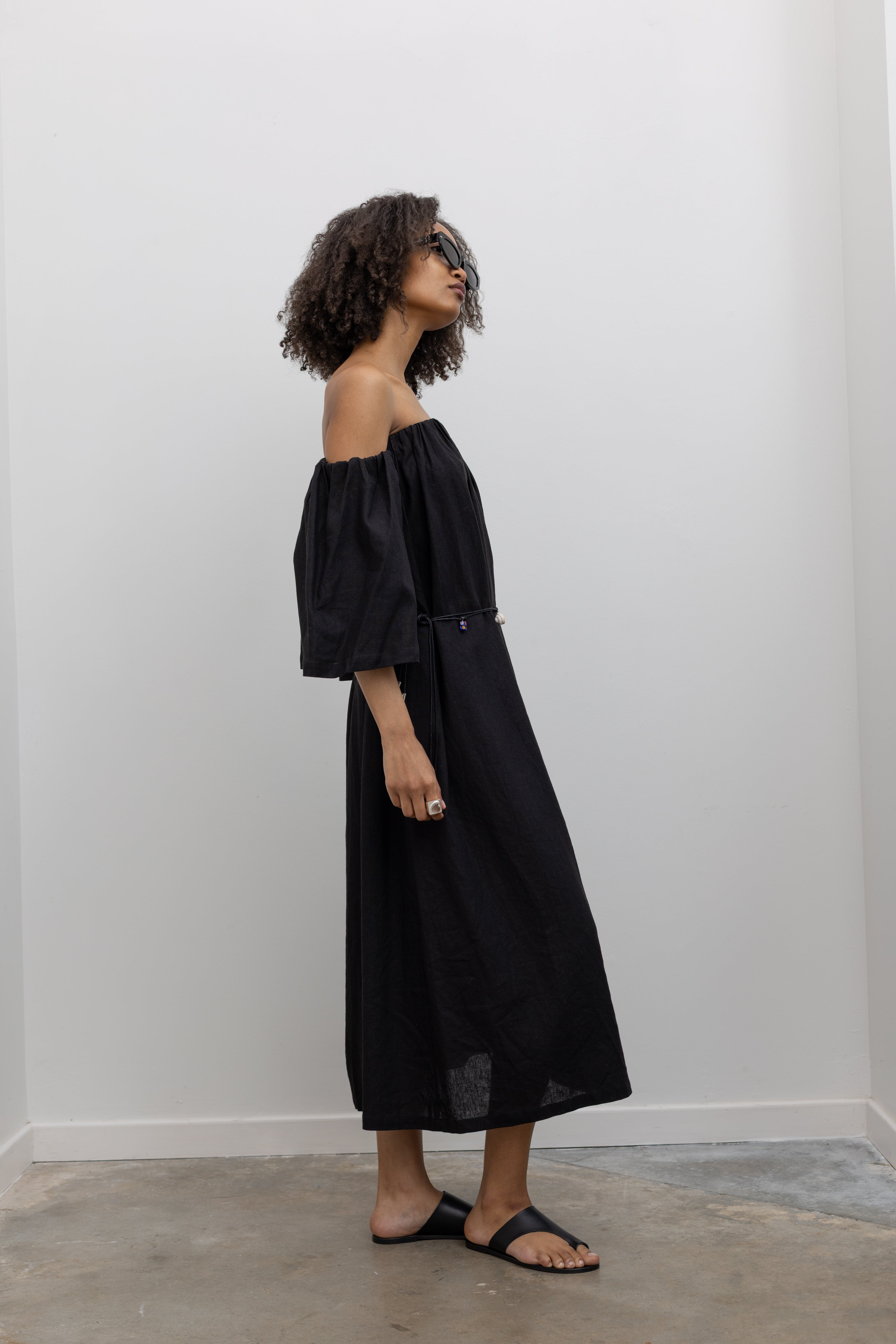 Side view of a woman in motion wearing chic summer fashion in a Alas Eius label black linen midi dress featuring removable sleeves paired with a beaded belt at an Alas Eius store.