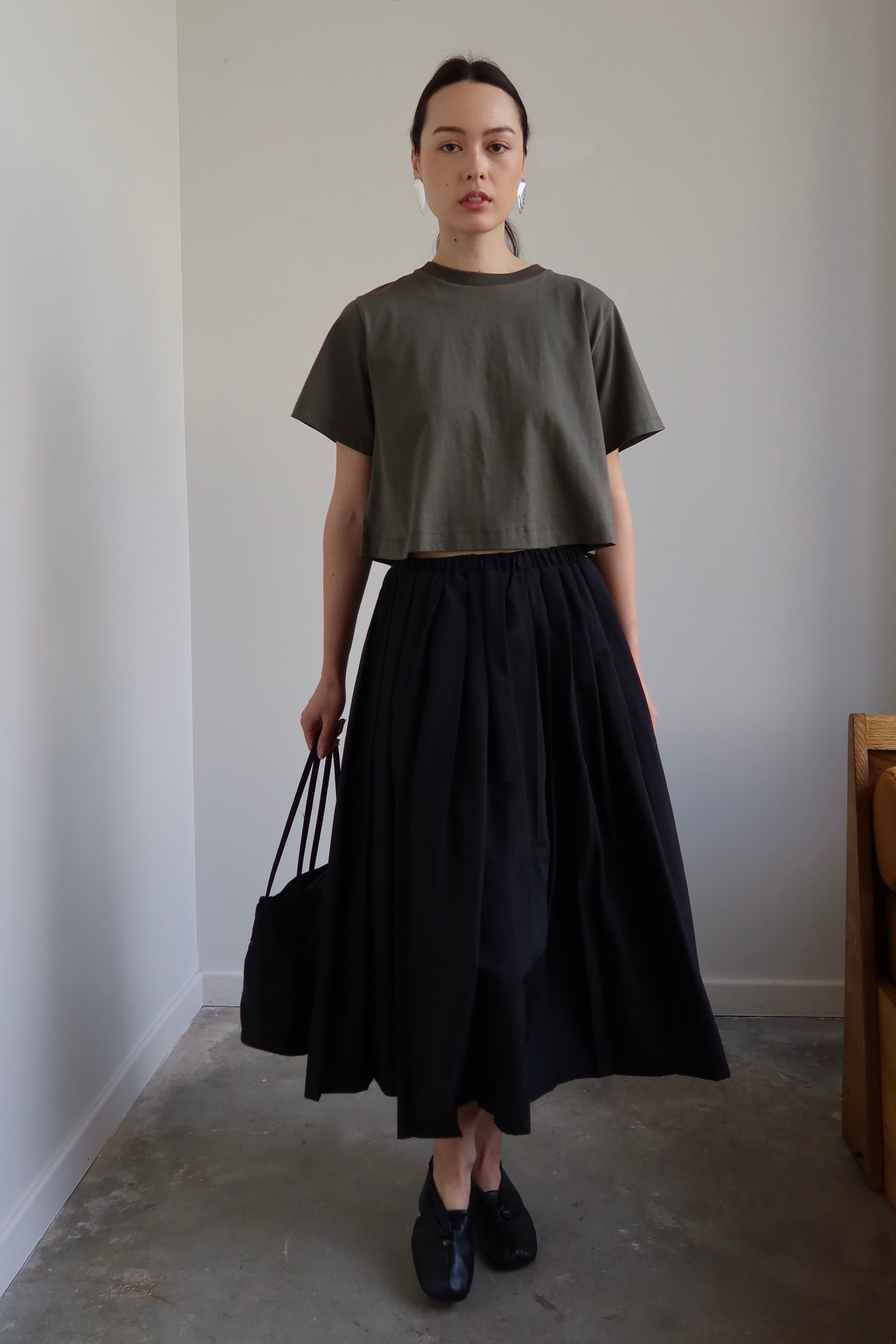 A person wearing a black pleated full skirt with an elastic waistband, paired with a grey t-shirt. The skirt has two side pockets and falls to the midi length.