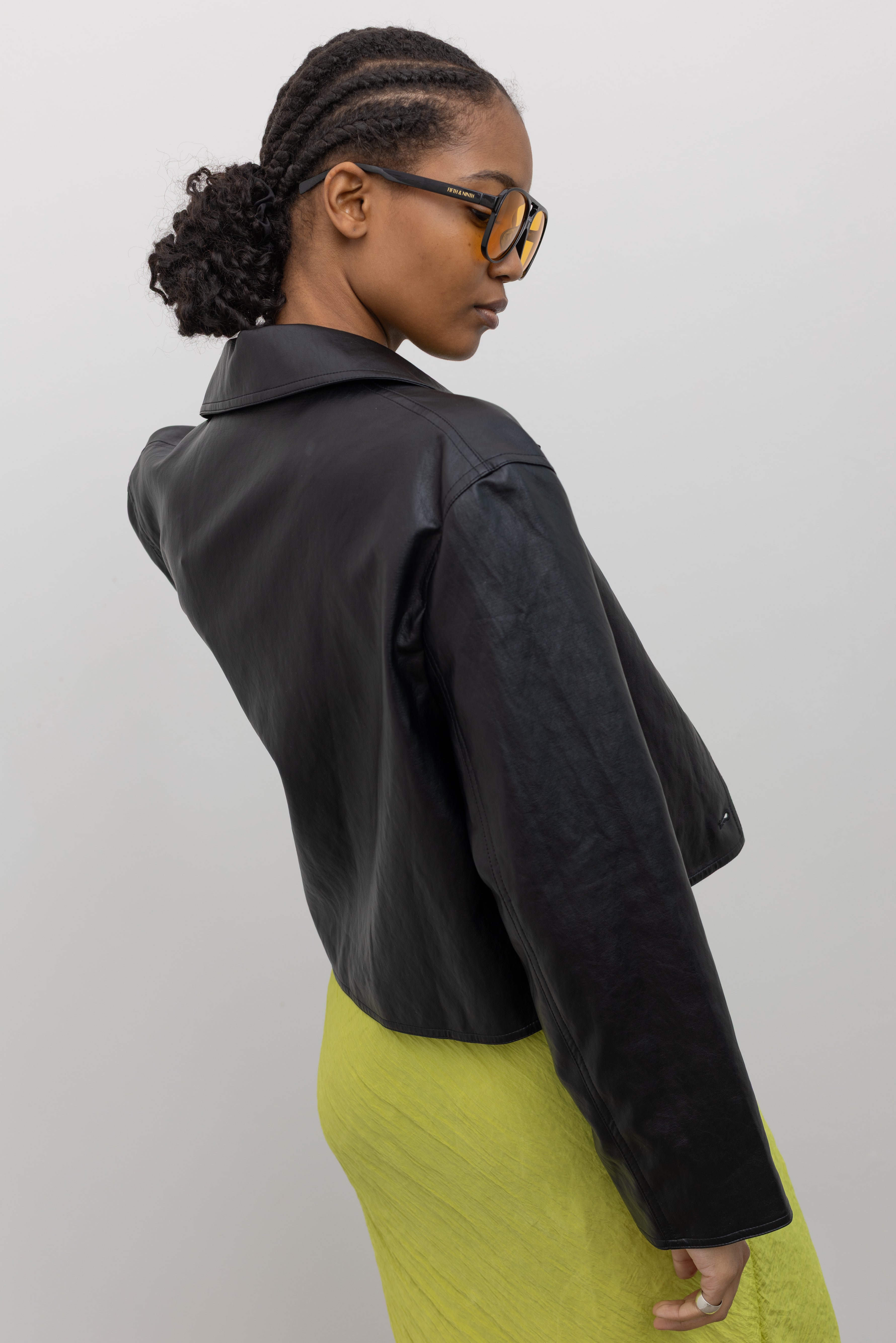 3/4 back view of a woman posing wearing chic and refined Alas Eius Label Black Cropped Vegan Leather Collared Jacket with a chartreuse dress at Alas Eius.  