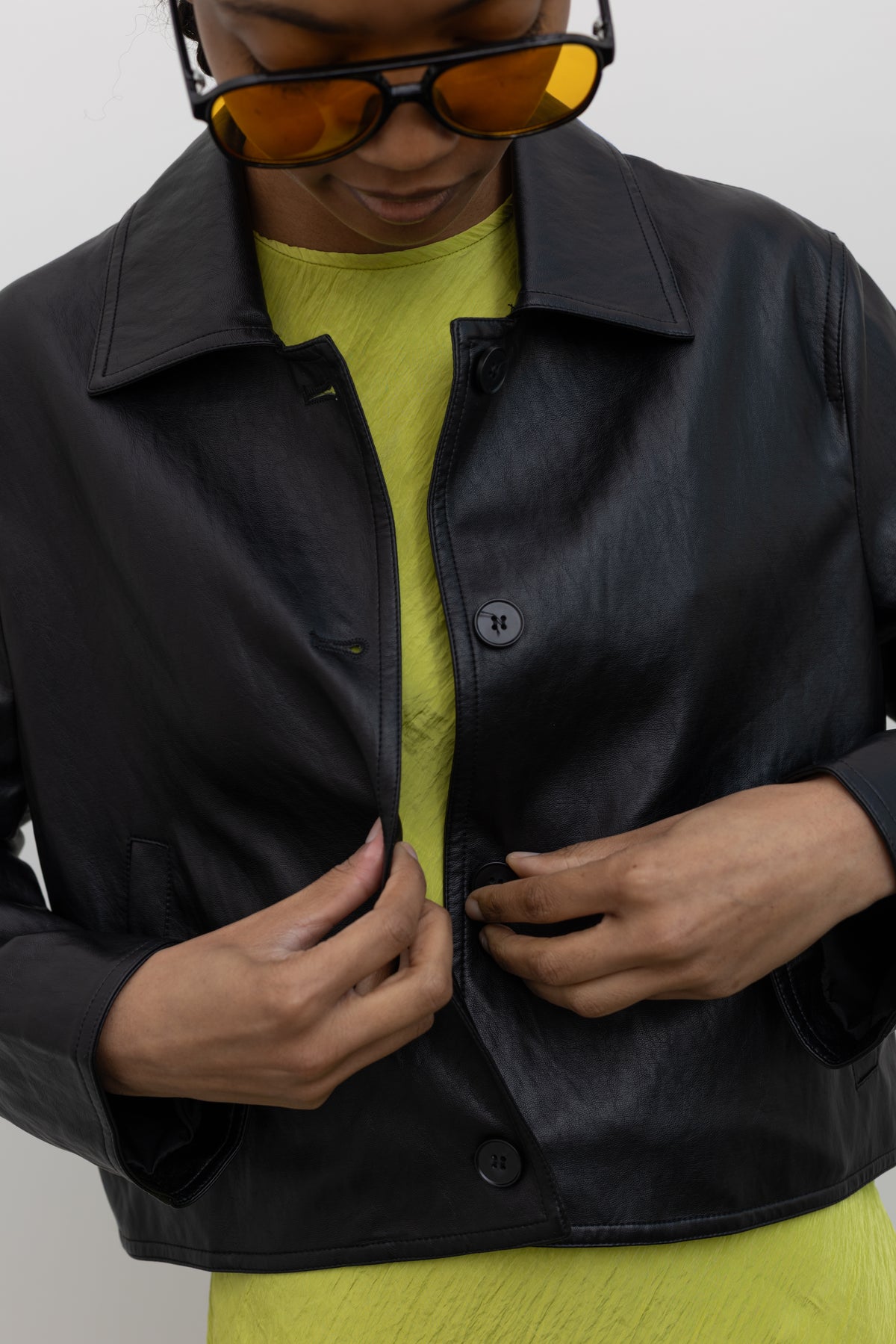 Close-up front view of a woman buttoning a versatile spring layering staple in a Alas Eius Label Black Cropped Vegan Leather Collared Jacket with a chartreuse dress at Alas Eius.  
