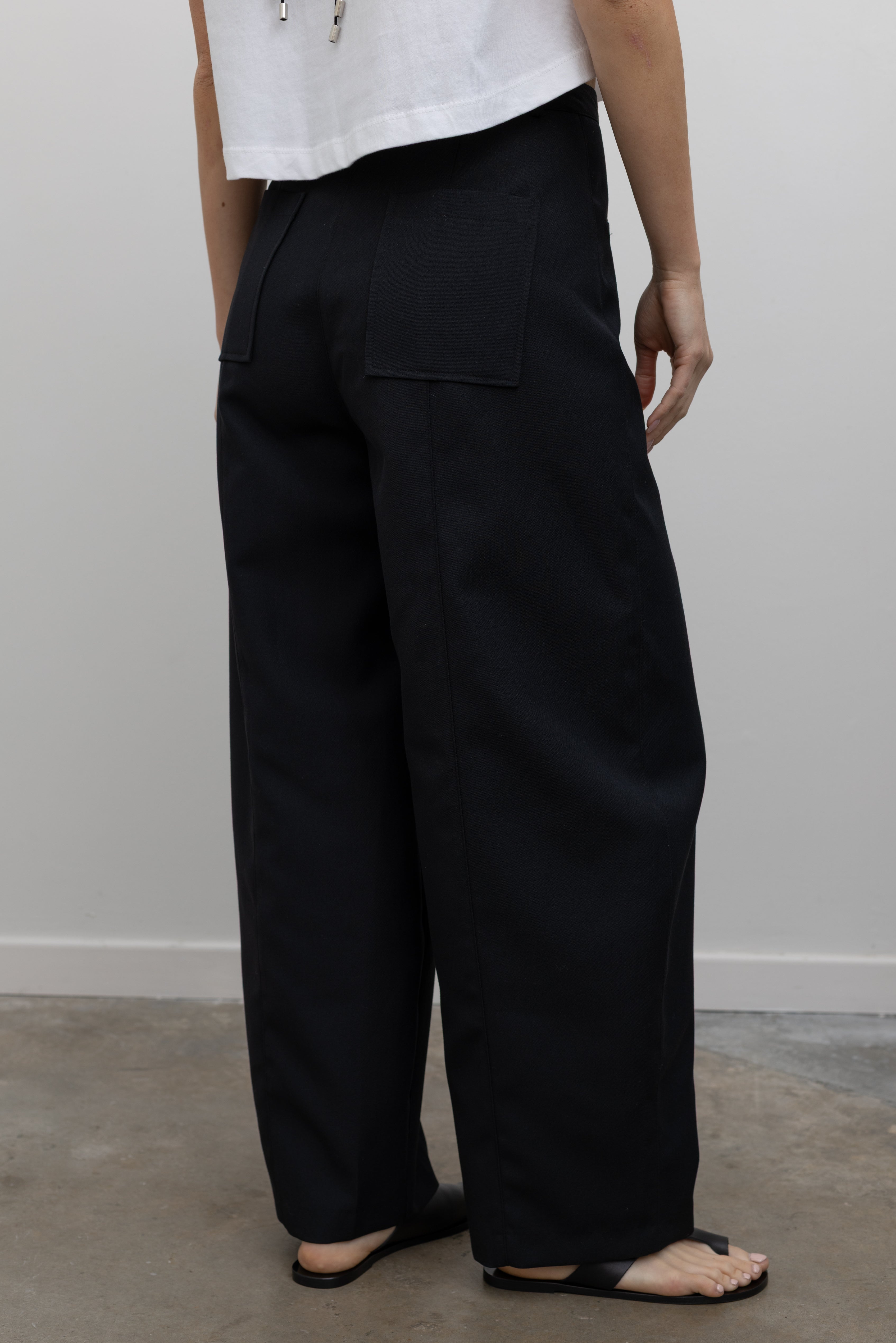 Back view of a woman standing wearing modern minimalist spring fashion in an Alas Eius black wool carrot high-waisted, wide-leg trouser paired with a white boxy cropped top at Alas Eius. 
