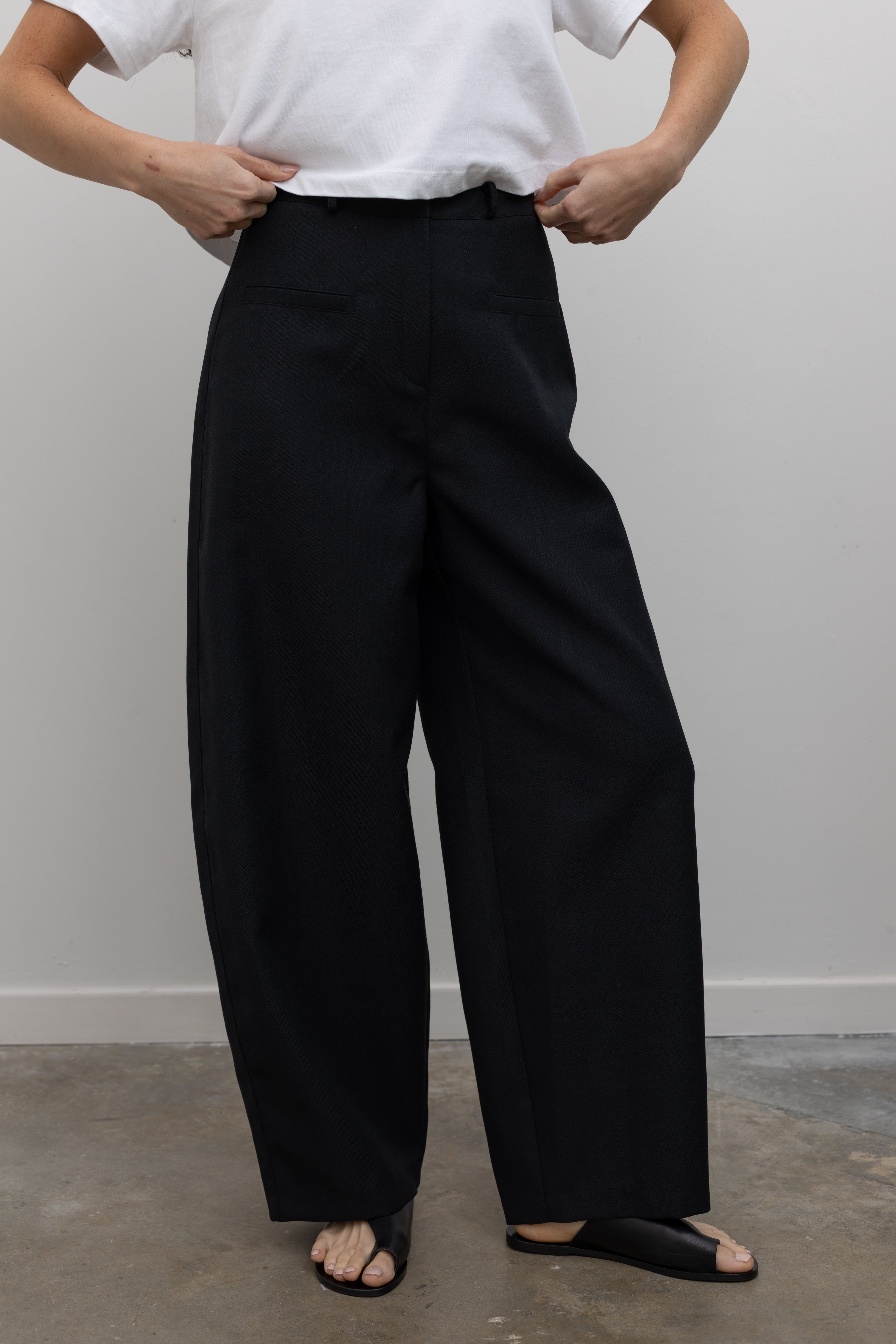 Front view of a woman standing wearing chic spring fashion in an Alas Eius black wool carrot high-waisted, wide-leg trouser paired with an Alas Eius white boxy cropped top at Alas Eius. 