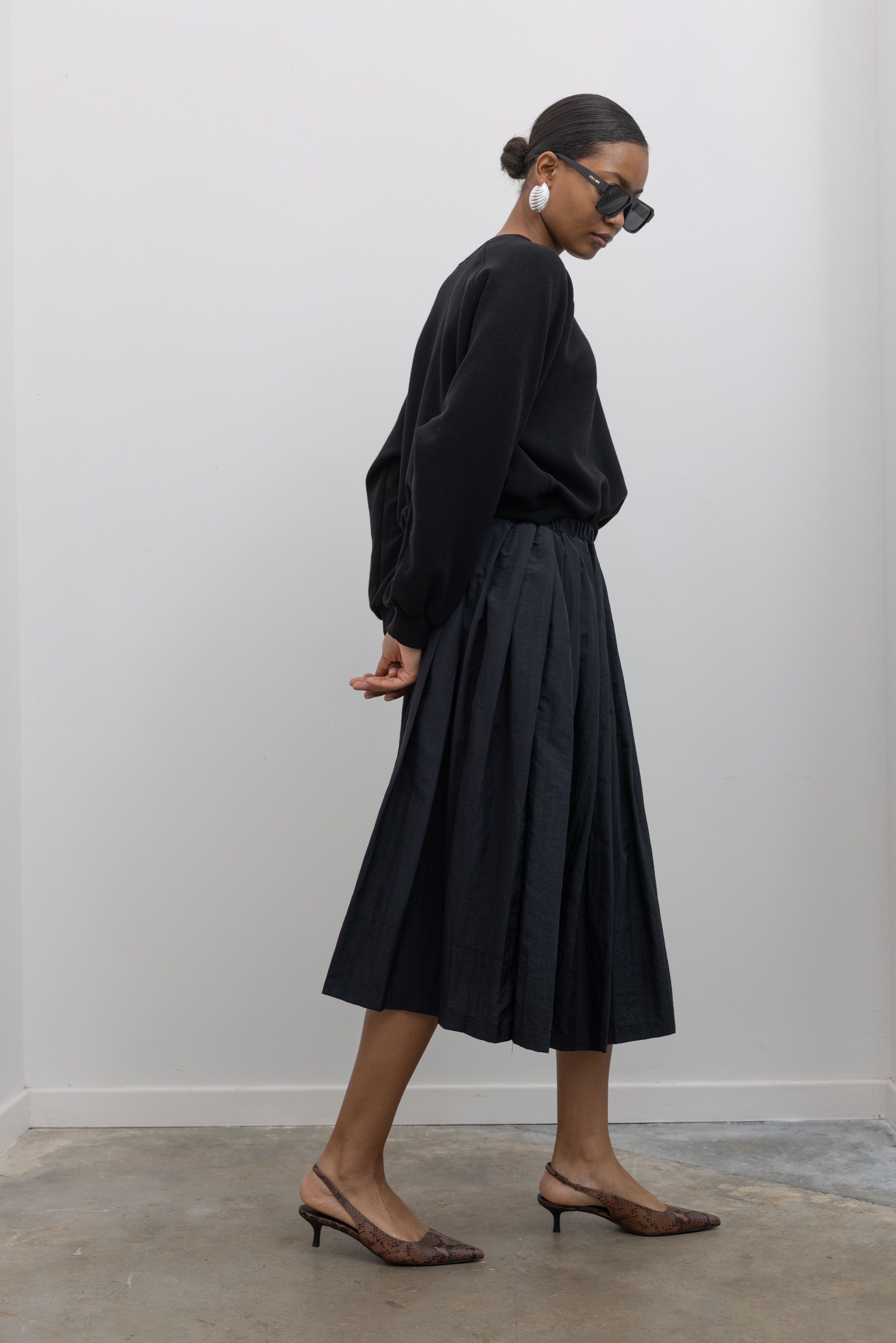 Side view of a woman in motion posing wearing an elegant outfit including a sophisticated Alas Eius Label black lightweight nylon pleated midi skirt, featuring a fully lined silhouette and finished with an elastic waistband and convenient side pockets. 