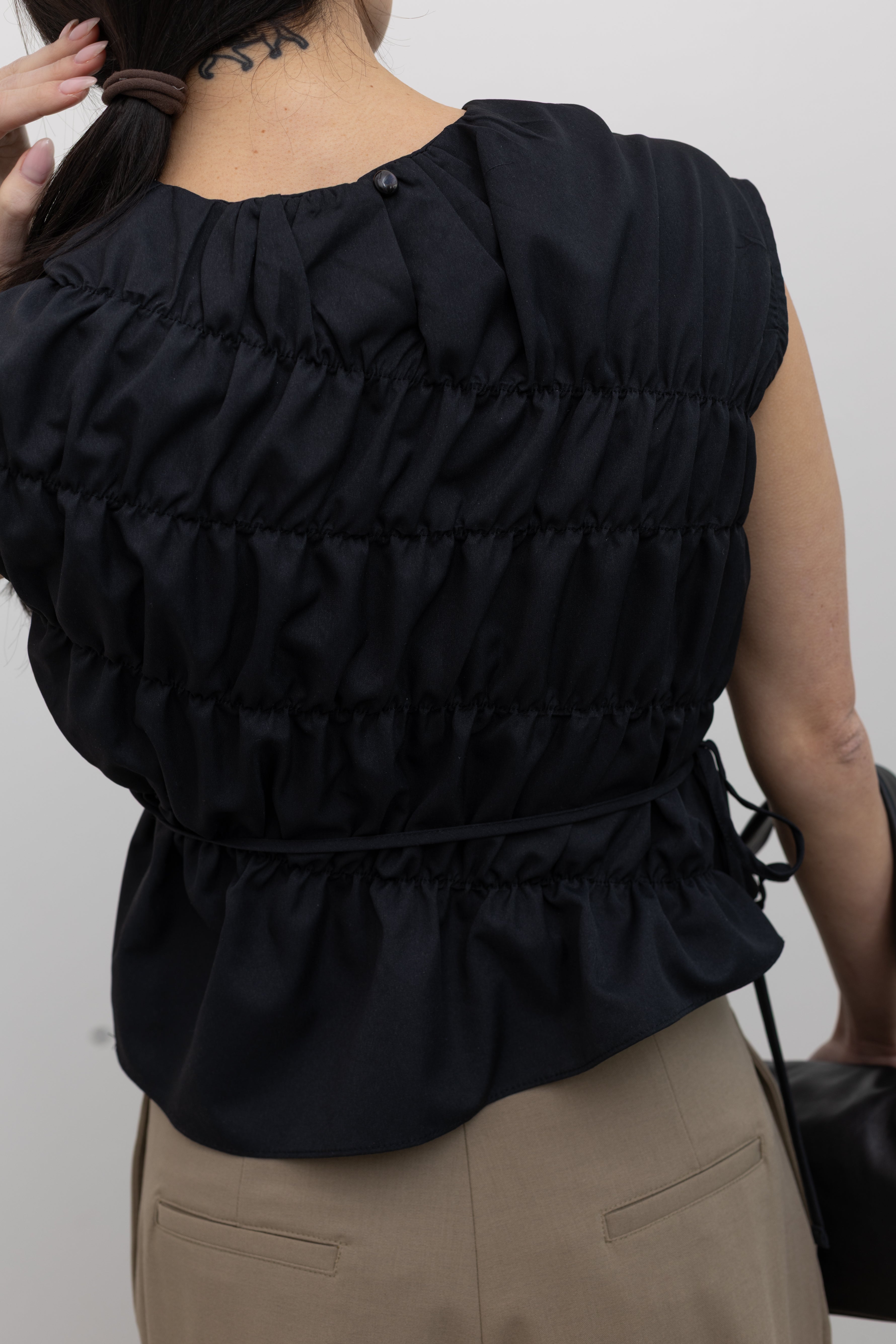 Close-up back view of a woman posing wearing contemporary and chic women's fashion in an Alas Eius Black Asymmetrical Cinched Top featuring a crew neckline, cap sleeves, and an optional waist tie at an Alas Eius store. 