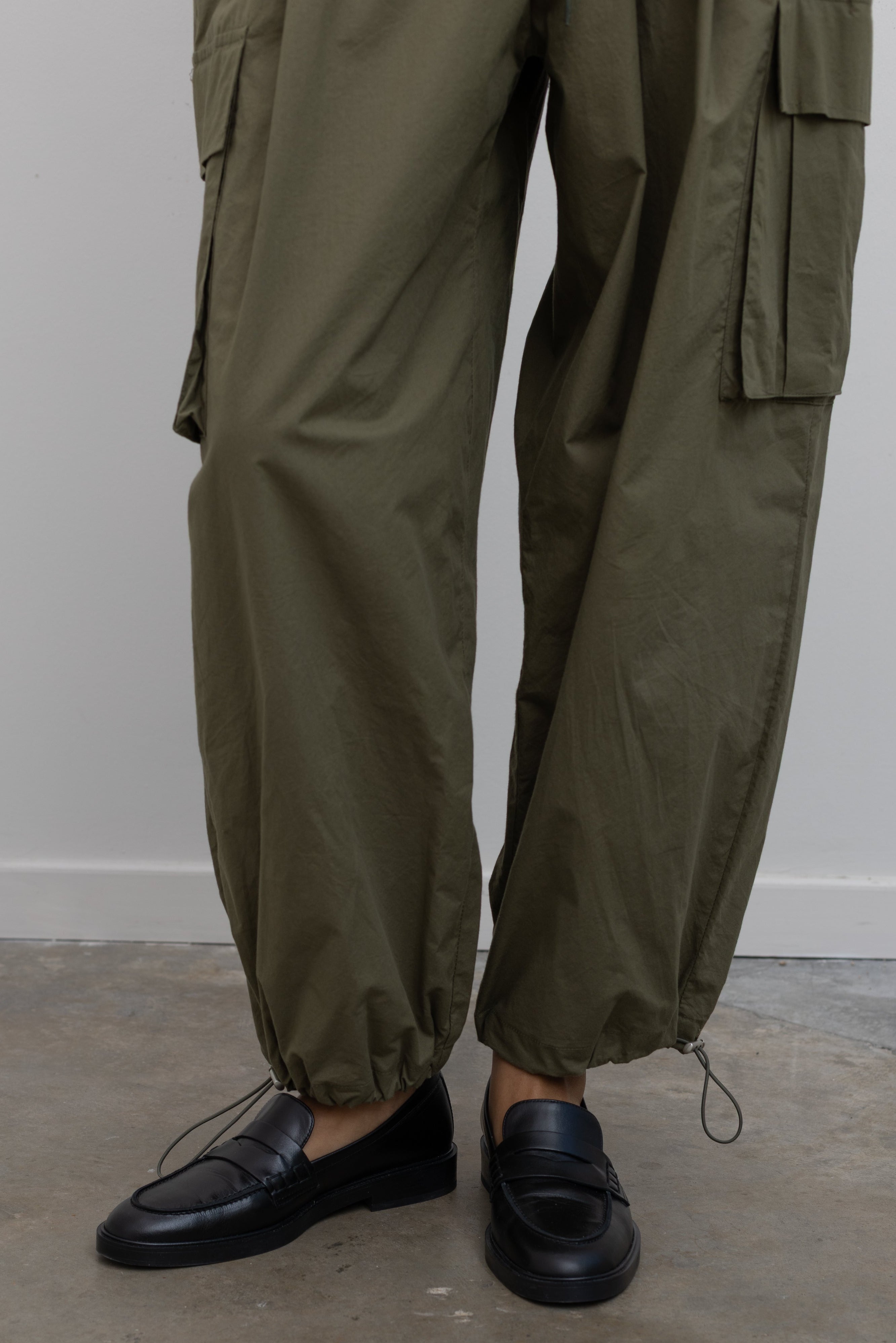 Woman standing wearing everyday fashion in a pair of Alas Eius brand khaki green lightweight cotton wide-leg cargo pants with drawstring ankles and a tie waist and extra pockets at Alas Eius 