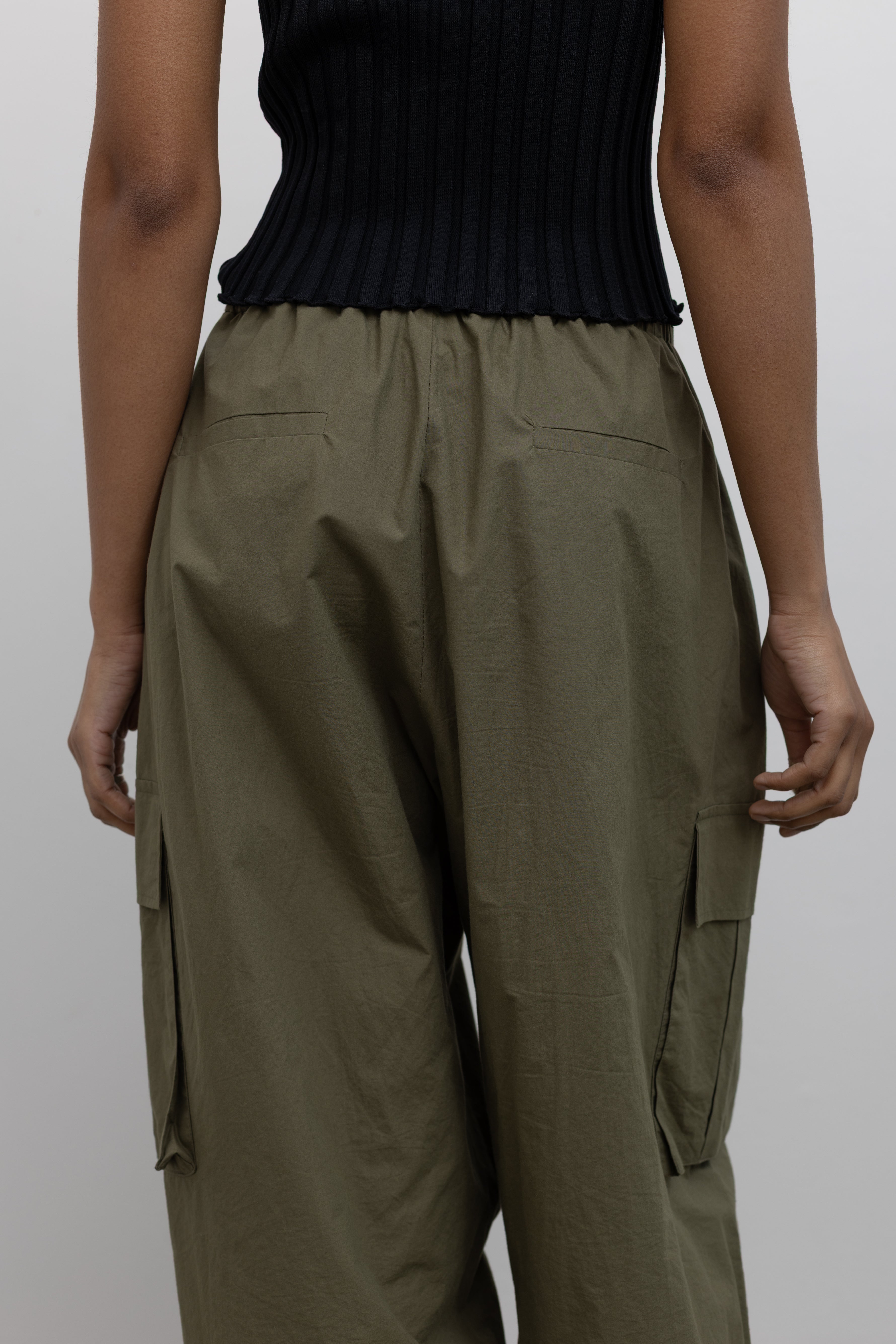 Back product view of Woman standing wearing everyday fashion in a pair of Alas Eius brand khaki green lightweight cotton wide-leg cargo pants with drawstring ankles and a tie waist and extra pockets at Alas Eius 
