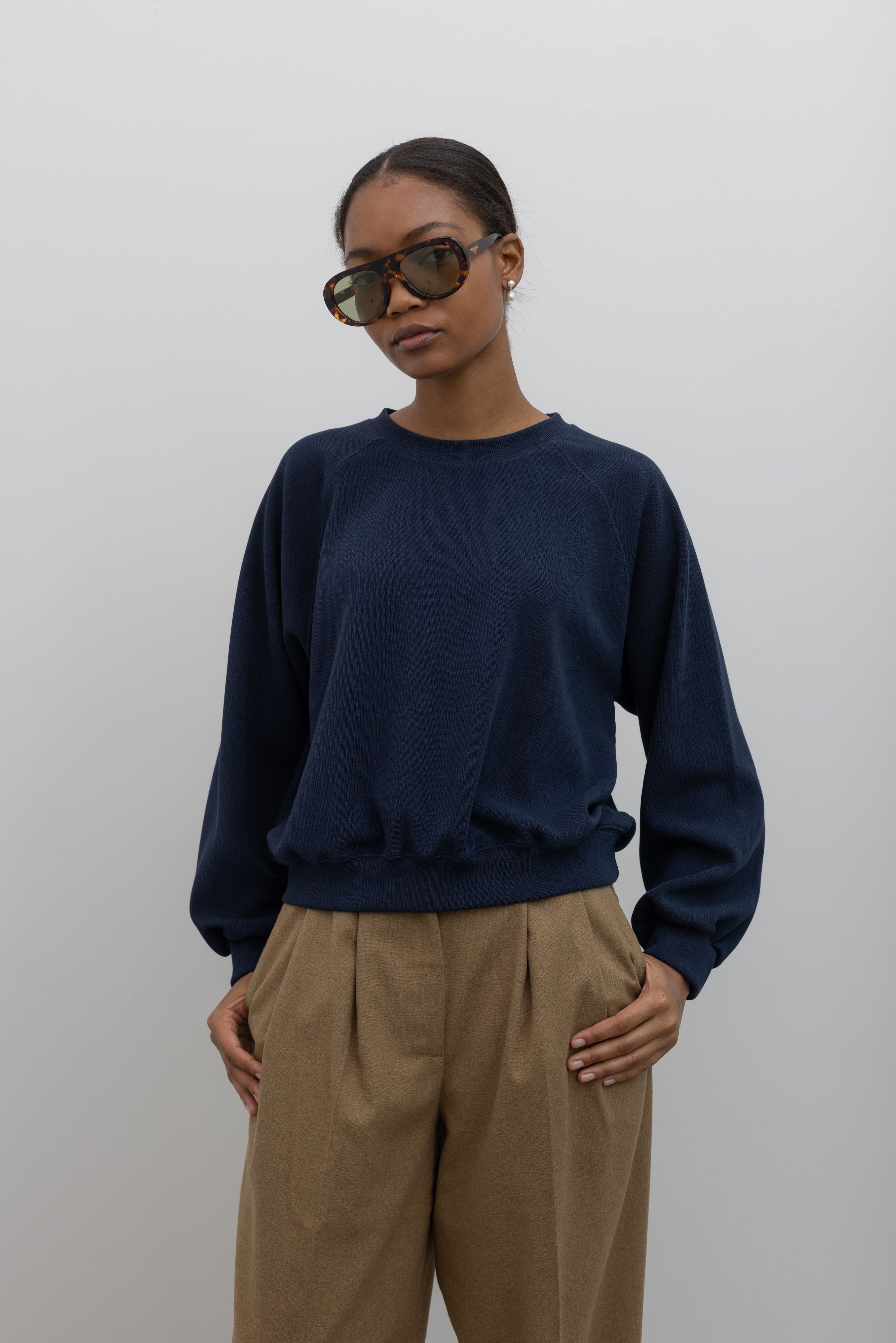 Front view of a woman standing wearing a capsule wardrobe essential style in an Alas Eius label navy blue raglan sleeve elevated sweatshirt with a slightly cropped hem, landing at the waistband. Paired with tan trousers and sunglasses at Alas Eius. 