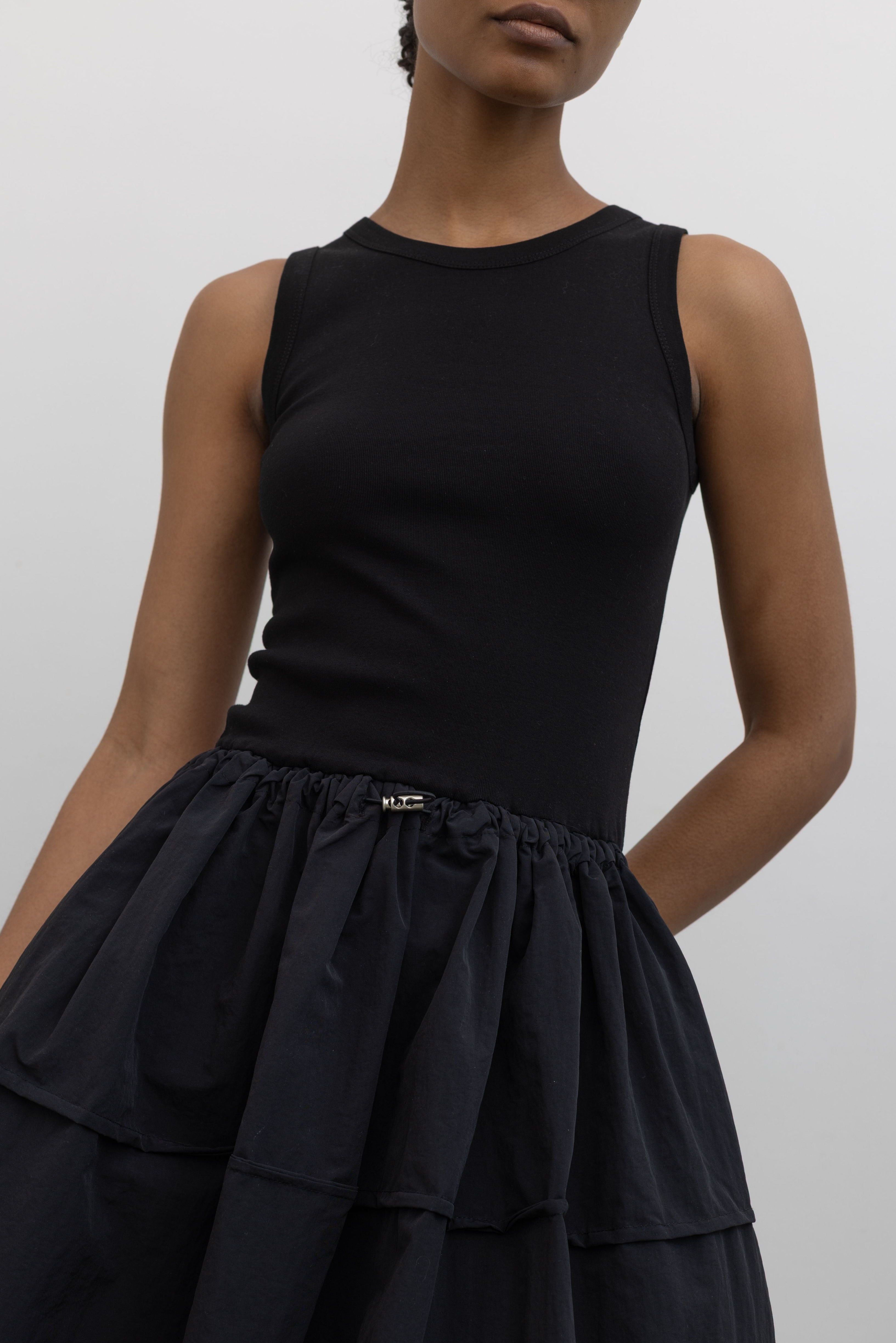 Woman wearing Spring fashion in a flattering Alas Eius brand sleeveless dress with a cotton bodice and a structured nylon skirt cinched by an adjustable drawstring waist paired with black Salomon sneakers and sunglasses at Alas Eius 