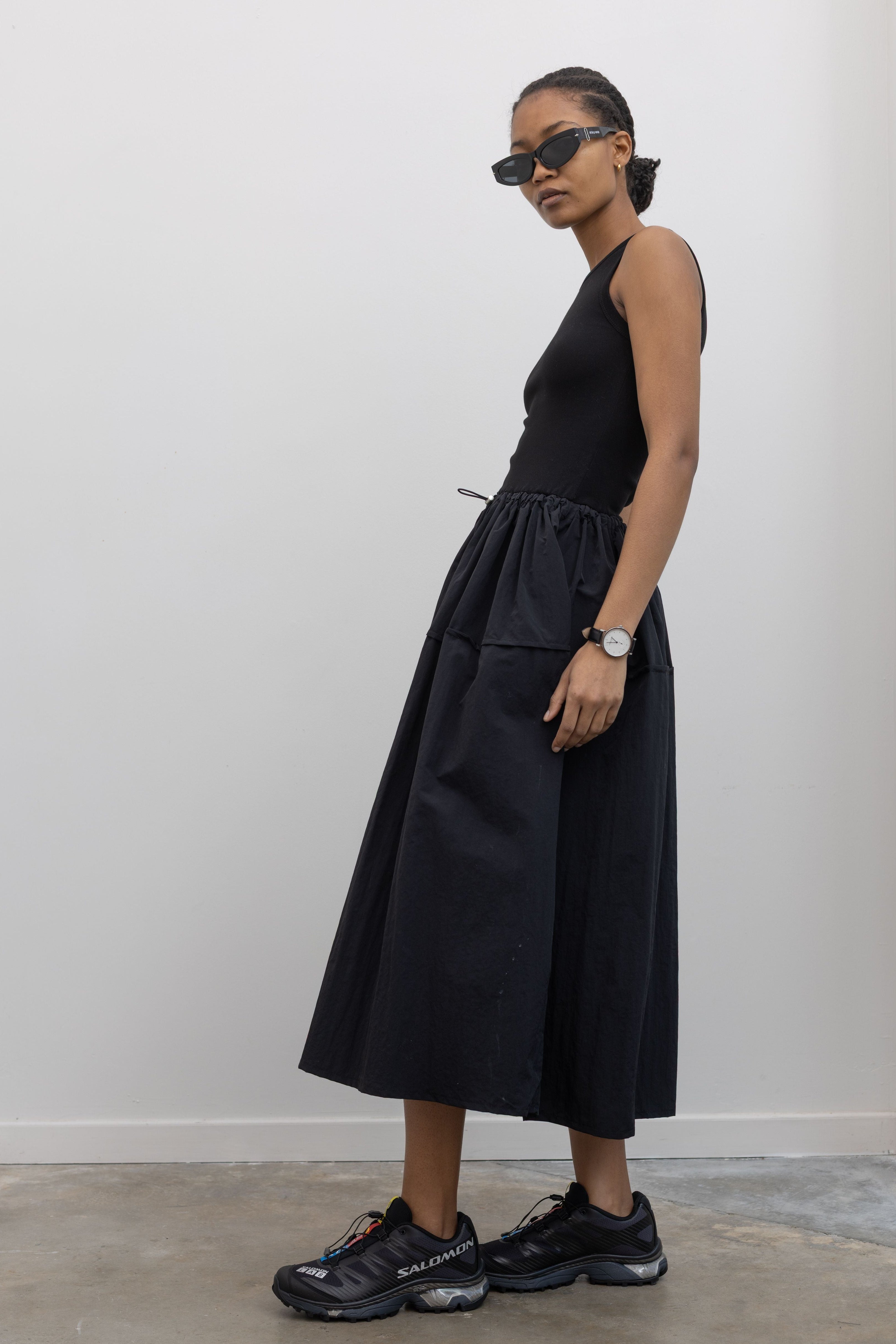 Side view of a Woman wearing Spring fashion in a flattering Alas Eius brand midi dress with a cotton bodice and a structured nylon skirt cinched by an adjustable drawstring waist paired with black Salomon sneakers and sunglasses at Alas Eius 