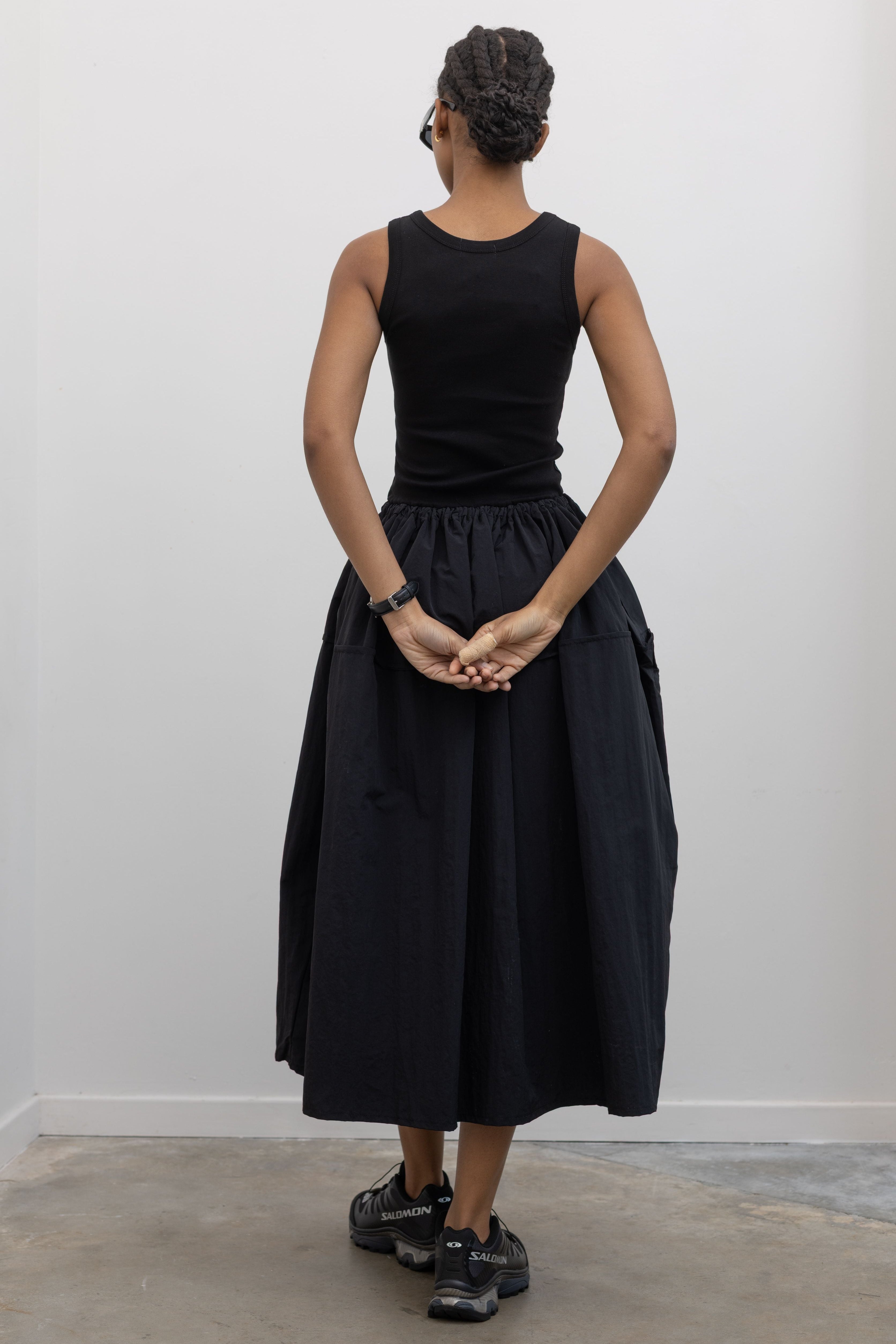 Back view of a Woman wearing Spring fashion in a flattering Alas Eius brand midi dress with a cotton bodice and a structured nylon skirt cinched by an adjustable drawstring waist paired with black Salomon sneakers and sunglasses at Alas Eius 