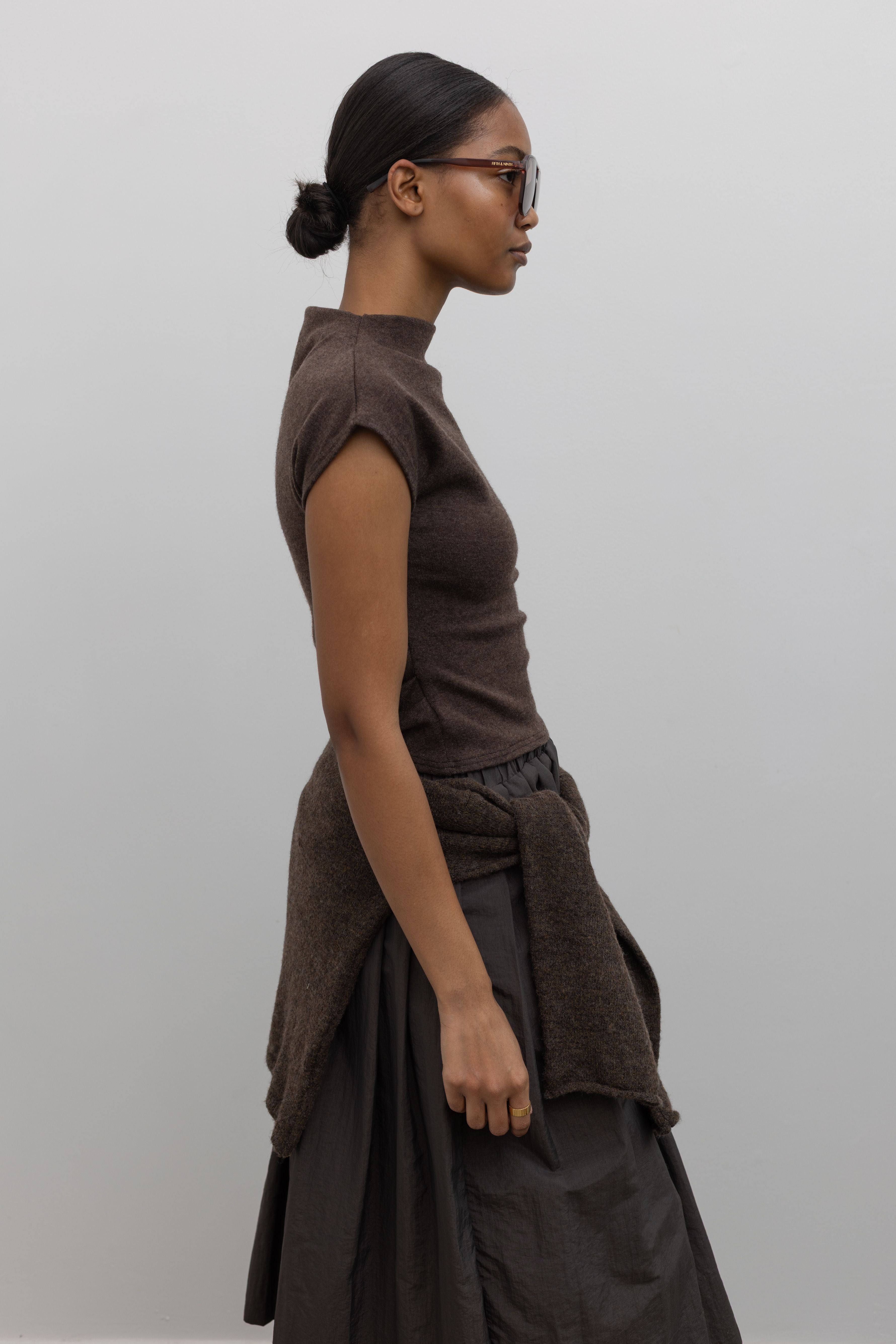 Side view of a woman wearing modern minimalist fashion in an Alas Eius label brown mock neck knit slim fit top with flattering cap sleeves at Alas Eius