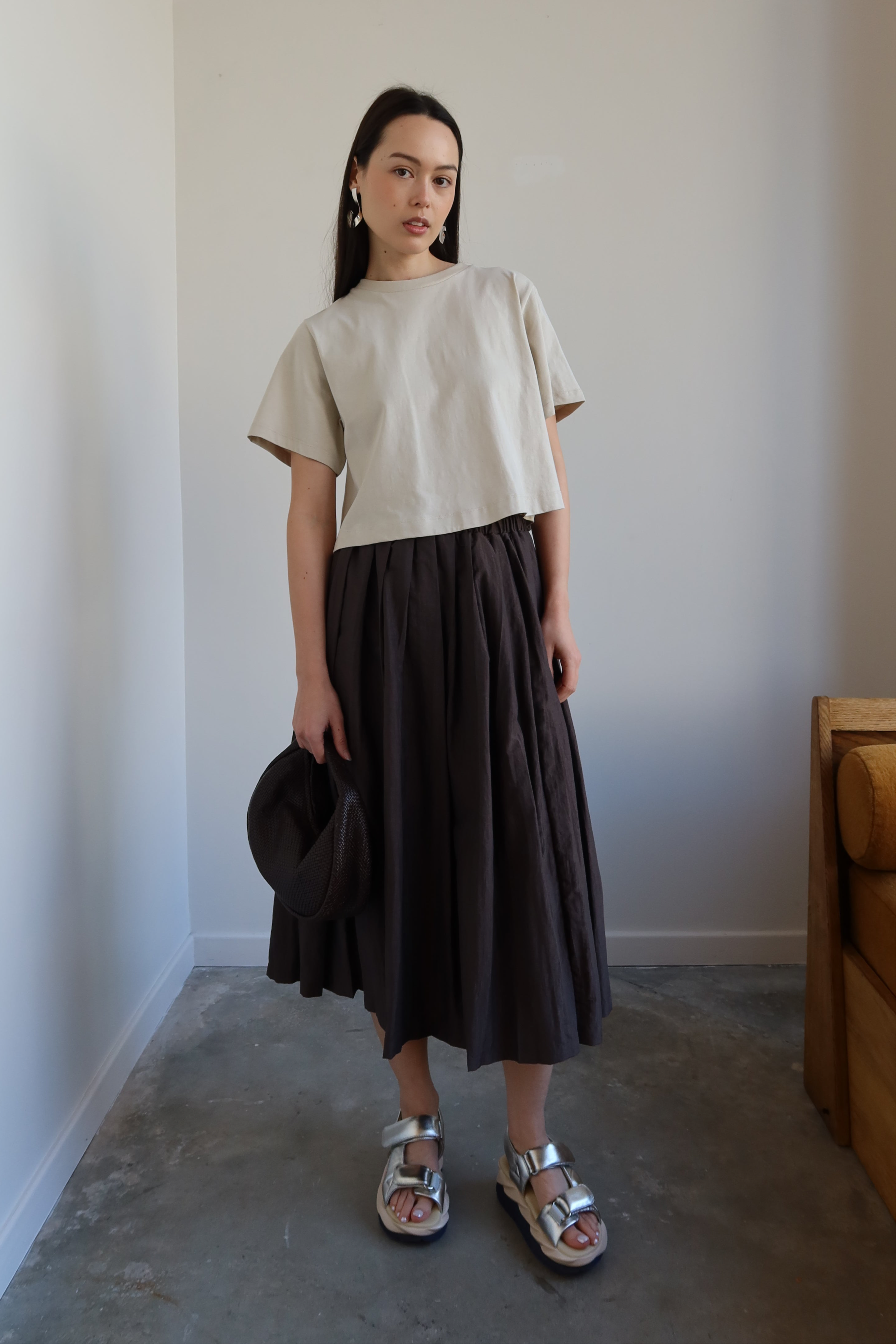 A person wearing a beige top and a brown pleated full skirt with side pockets, standing in a room with minimalist decor.