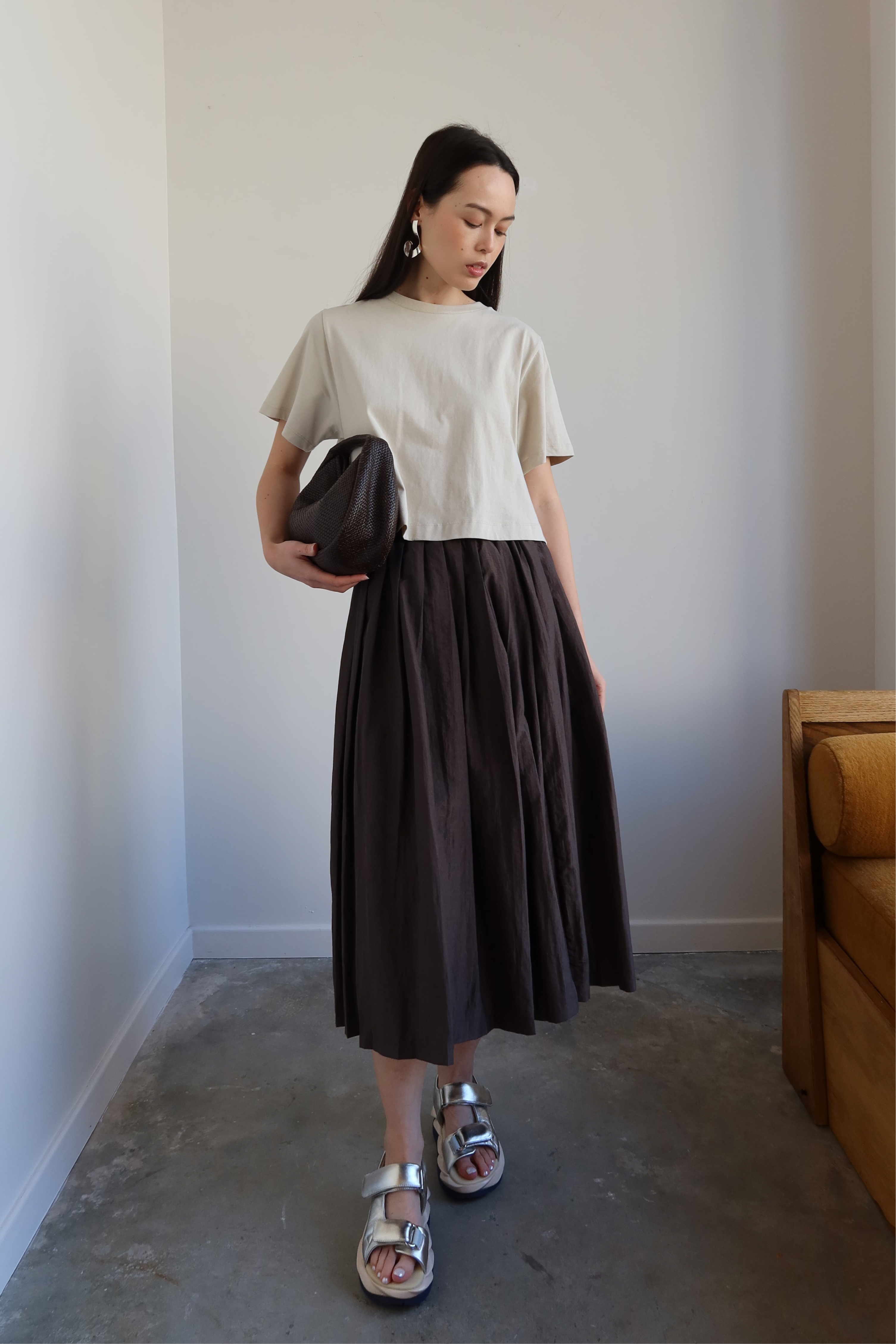 A person wearing a beige top and a brown pleated full skirt with side pockets, standing in a room with minimalist decor.