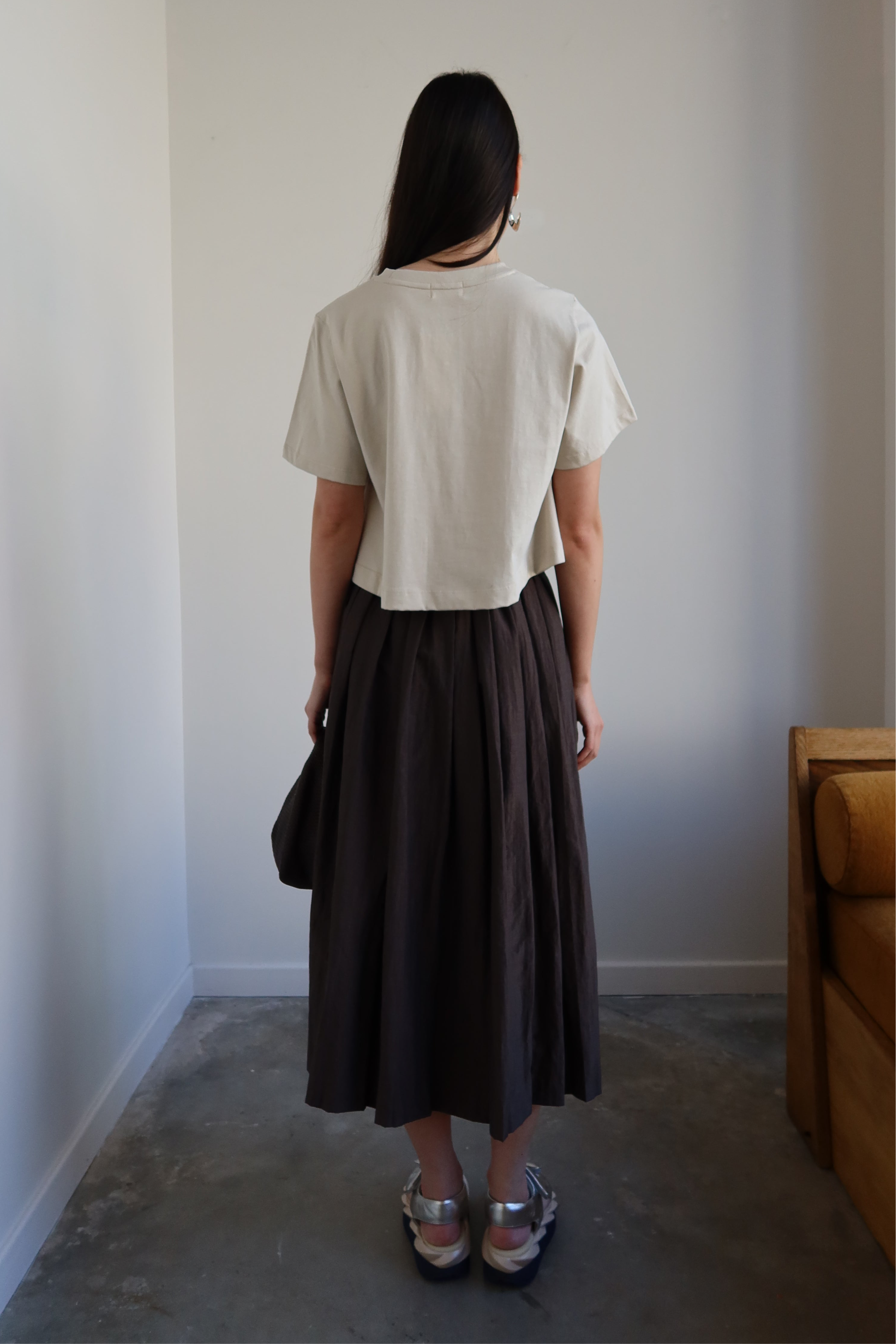 A person wearing a beige top and a brown pleated full skirt with side pockets, standing in a room with minimalist decor.