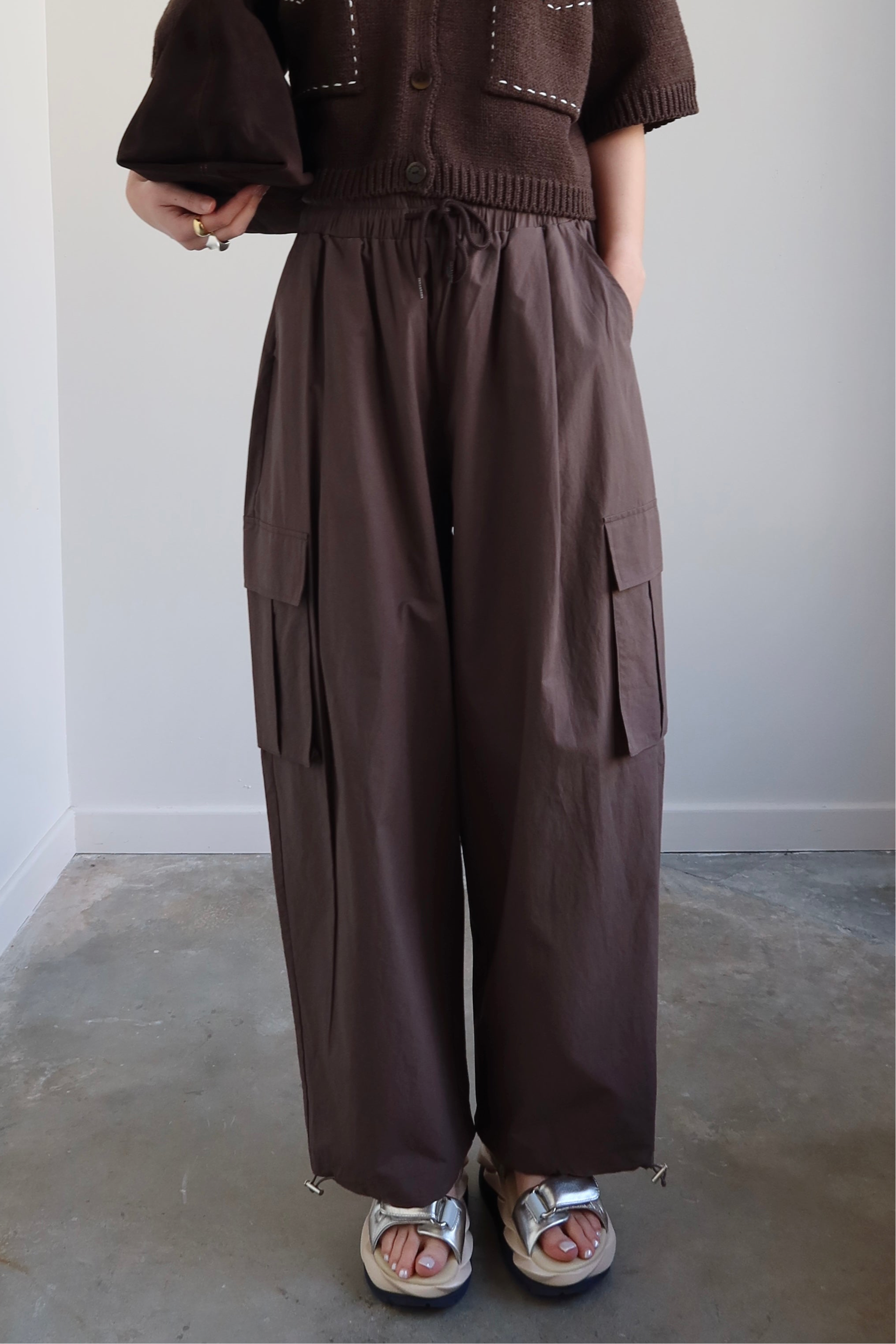 A person standing in a room wearing brown wide-leg cargo pants with a tie waist and drawstring ankles, paired with a brown top and white sandals.