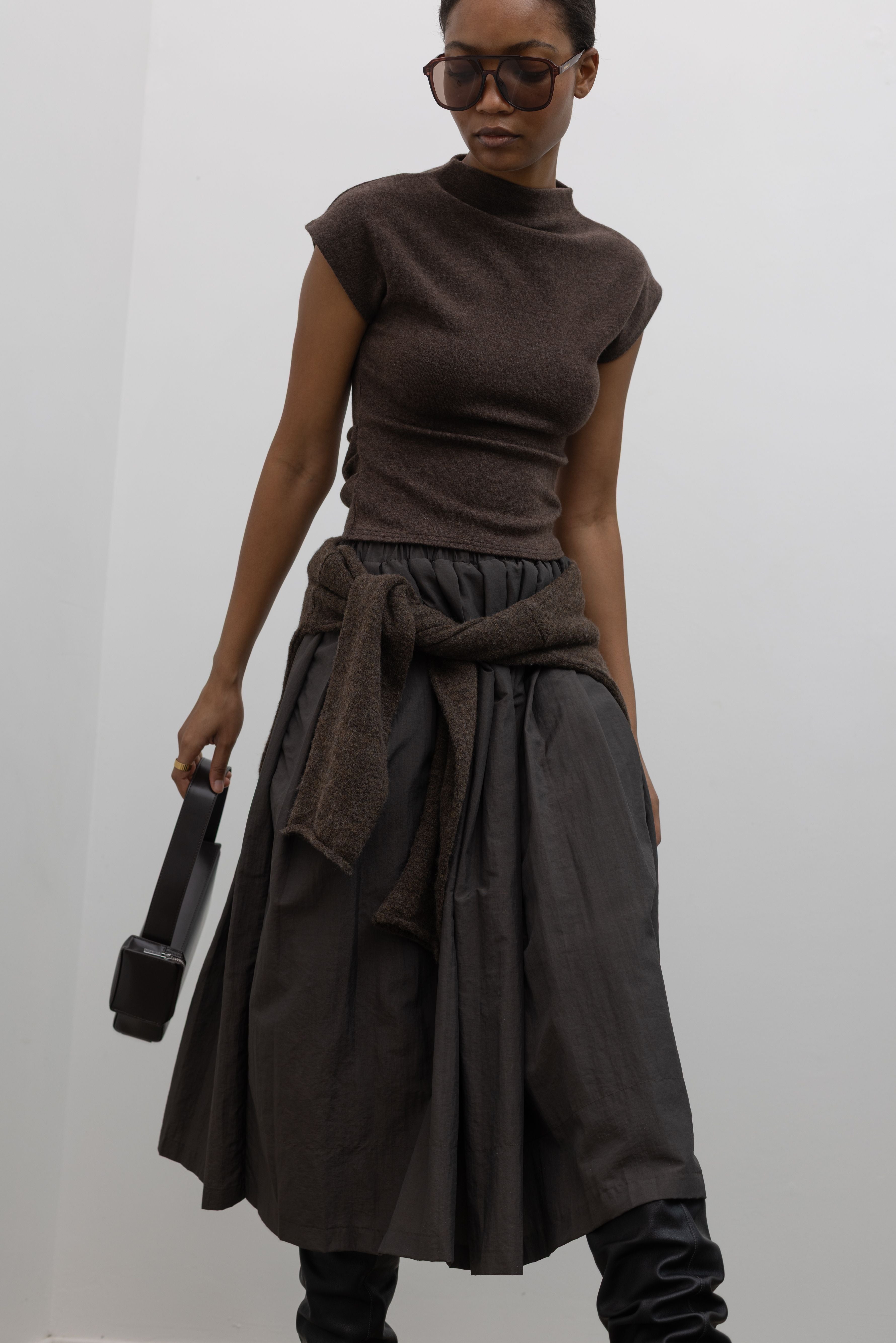 Front view of a woman wearing modest and chic fashion in an Alas Eius label brown mock neck knit slim fit top with flattering cap sleeves paired with an Alas Eius label nylon midi skirt and slouchy black leather boots. 