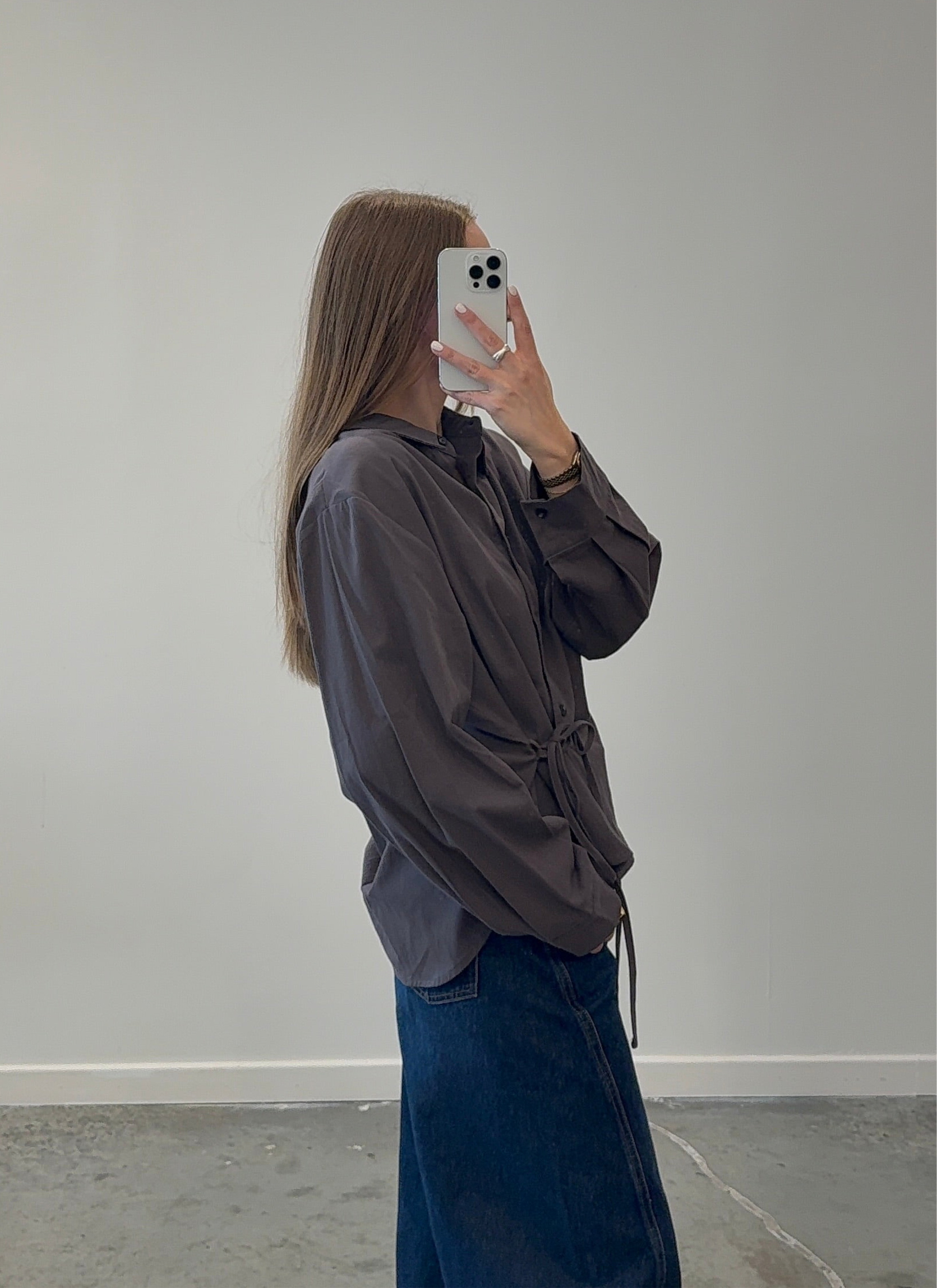 Person taking a mirror selfie wearing a brown jacket and blue jeans against a plain wall.