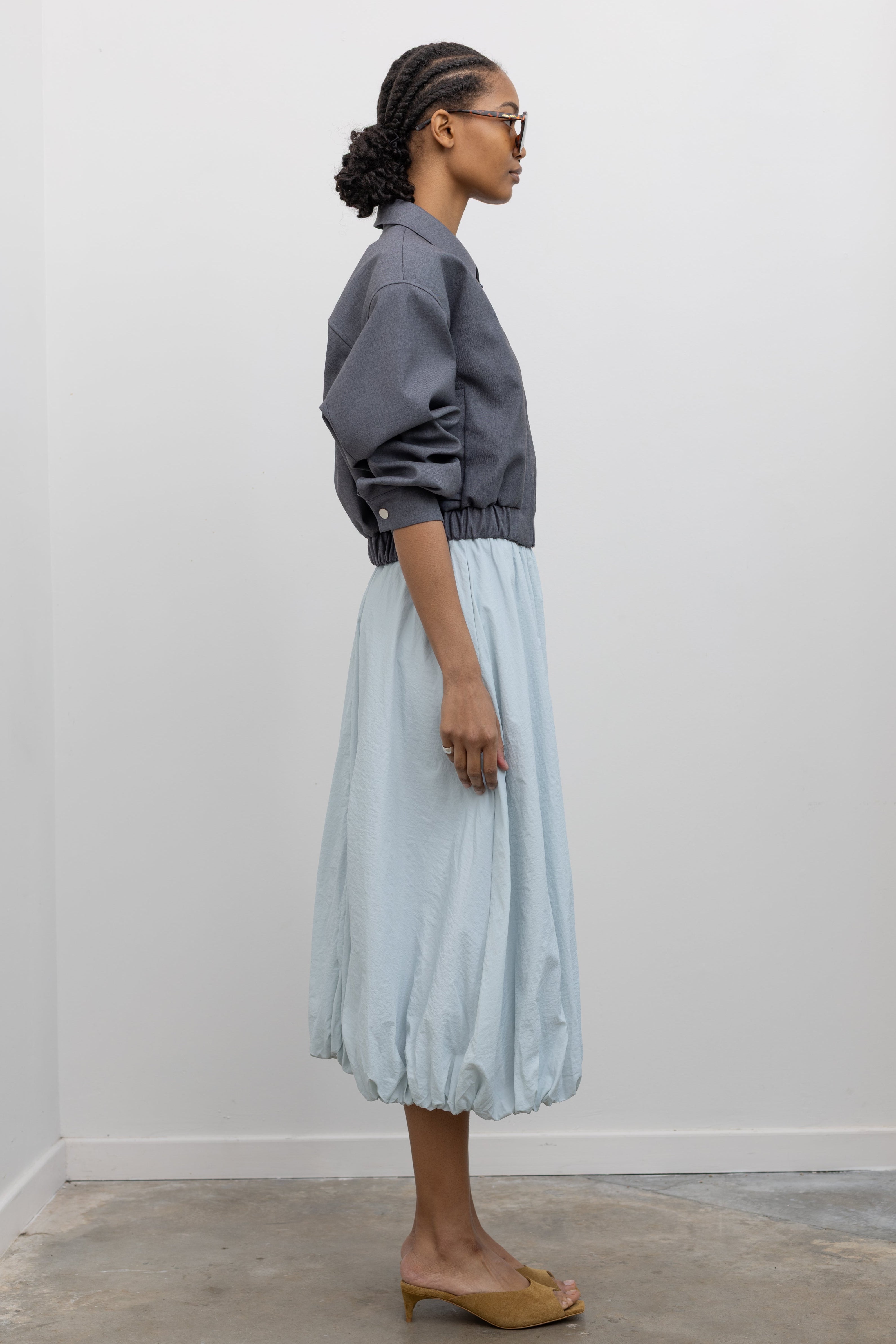 Side view of a woman wearing contemporary womenswear spring fashion in an Alas Eius label charcoal gray cropped tailored zip jacket featuring an elastic hem for a flattering cinched waist at an Alas Eius store. 