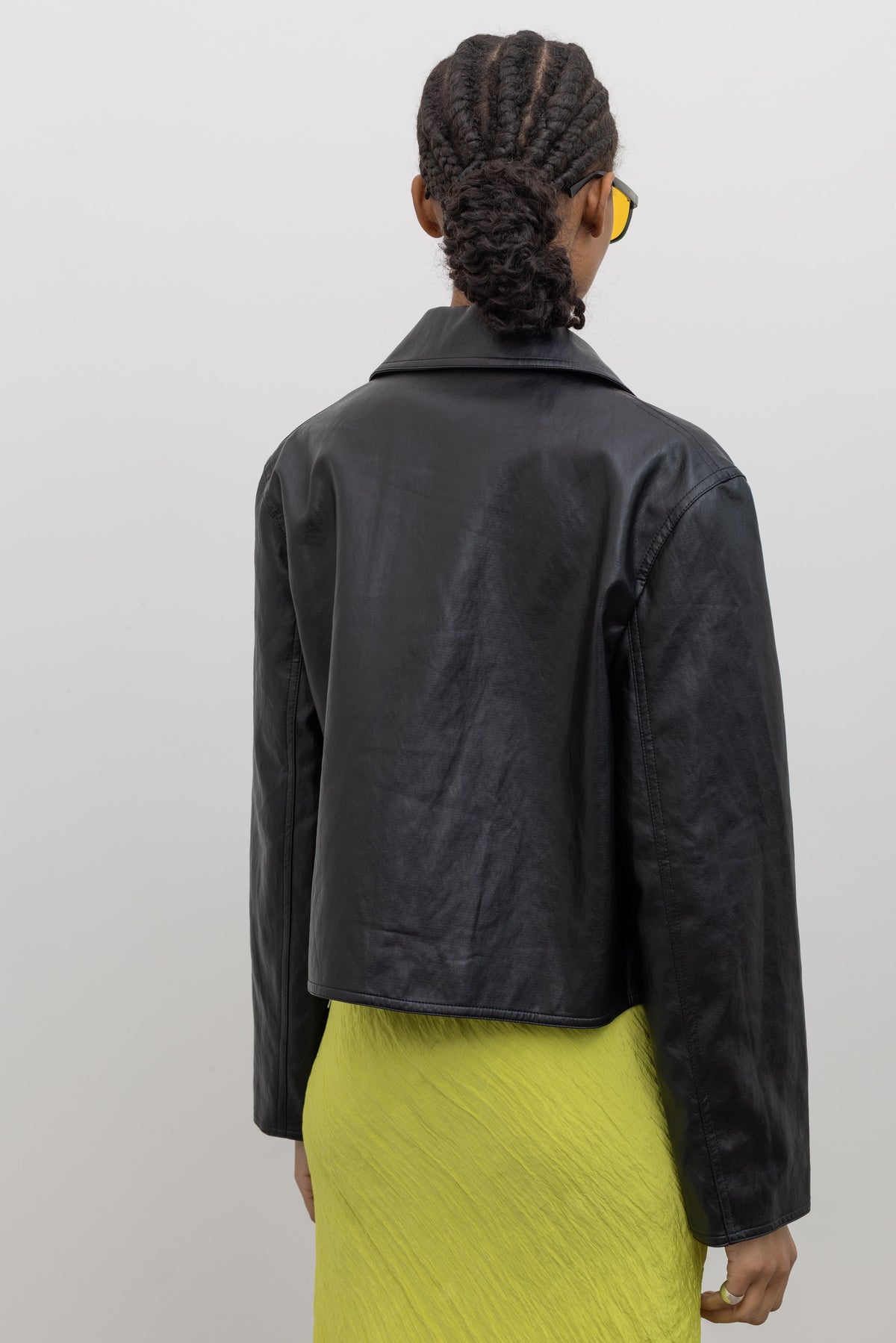 Back view of a woman posing wearing modern minimalist Alas Eius Label Black Cropped Vegan Leather Collared Button Front Jacket with a chartreuse dress at Alas Eius.  