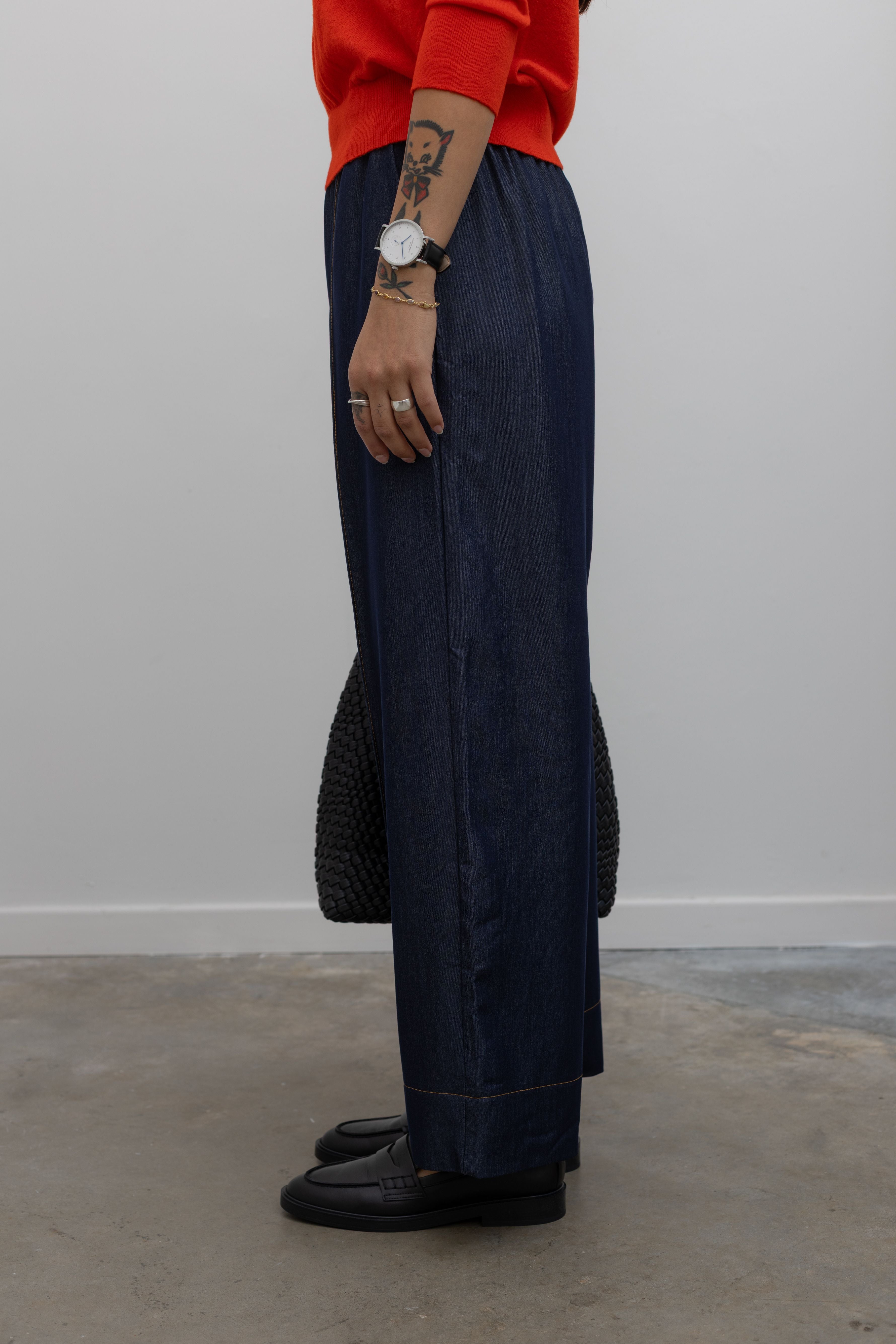 Side product view of Woman standing wearing an everyday outfit with a Tibi Red v-neck cashmere sweater and Alas Eius Label Tencel Blend Dark Wash Tailored Wide Leg Pull On Pants with black leather loafer and accessories at Alas Eius. 