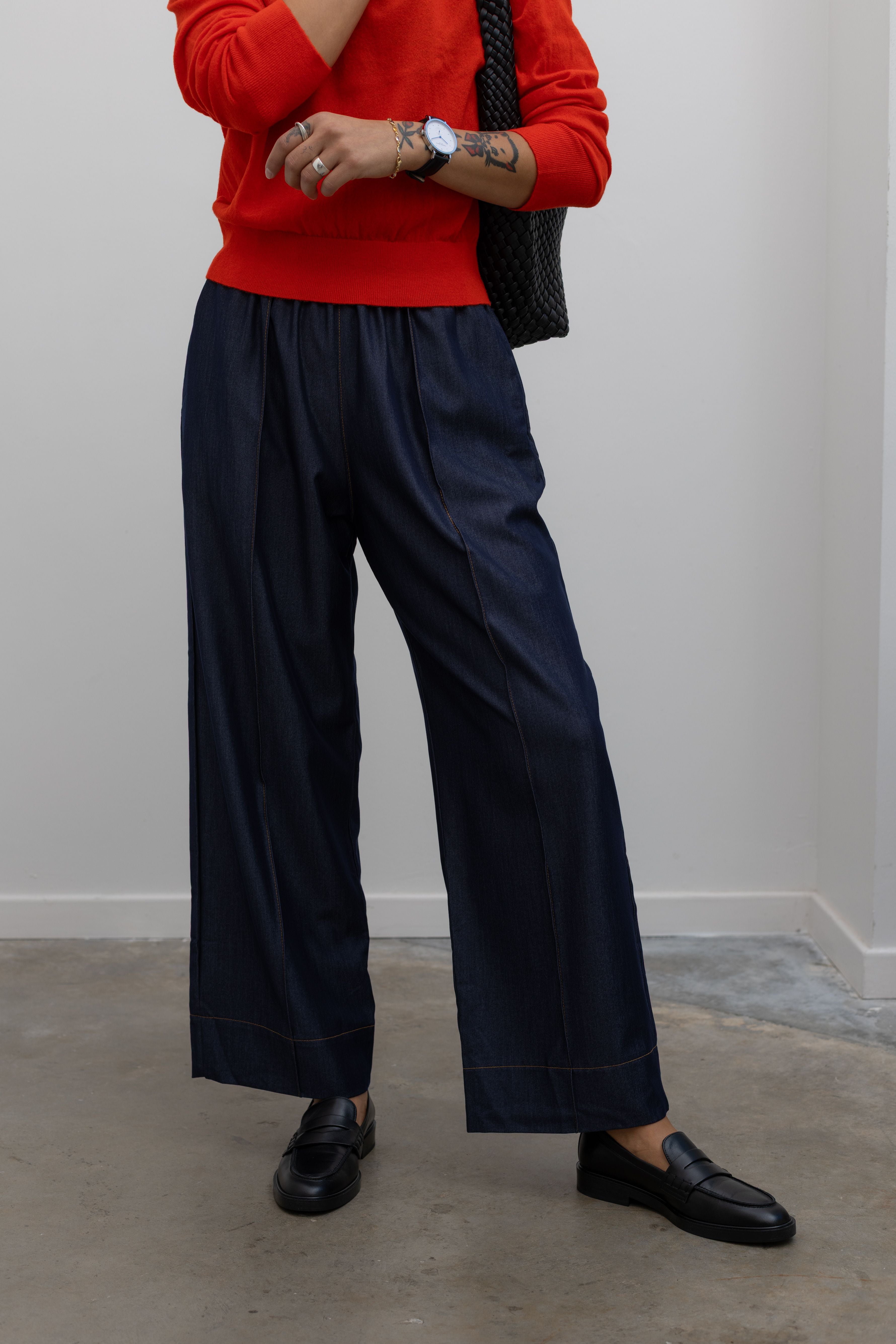 Front product view of Woman standing wearing an everyday outfit with a Tibi Red v-neck cashmere sweater and Alas Eius Label Tencel Blend Dark Wash Tailored Wide Leg Pull On Pants with black leather loafer and accessories at Alas Eius. 