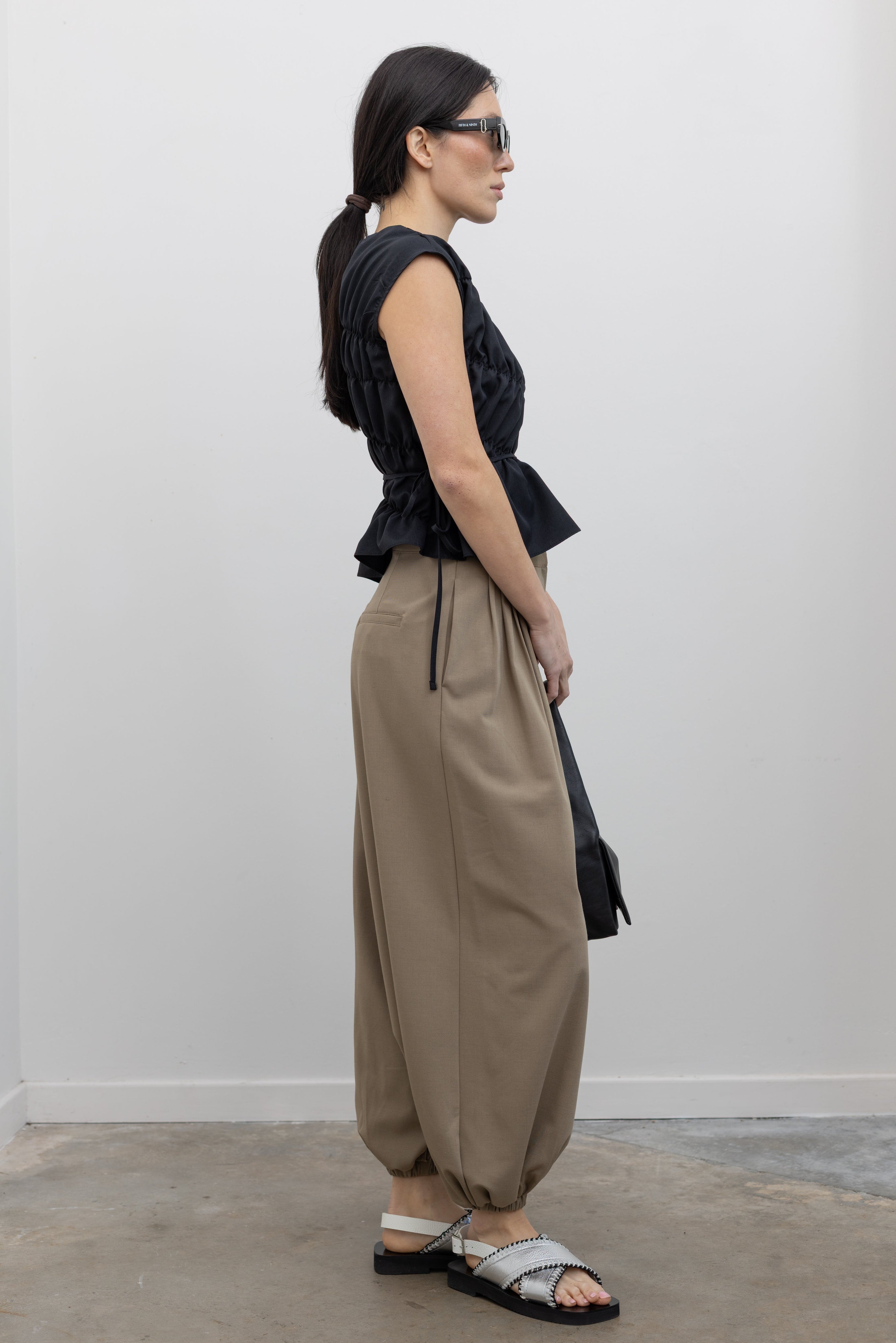 Side view of a woman posing wearing contemporary and chic women's fashion in an Alas Eius Black Asymmetrical Cinched Top featuring a crew neckline, cap sleeves, and an optional waist tie paired with elevated khaki cinched ankle pants, silver sandals, and a black leather St. Agni bag at an Alas Eius store. 