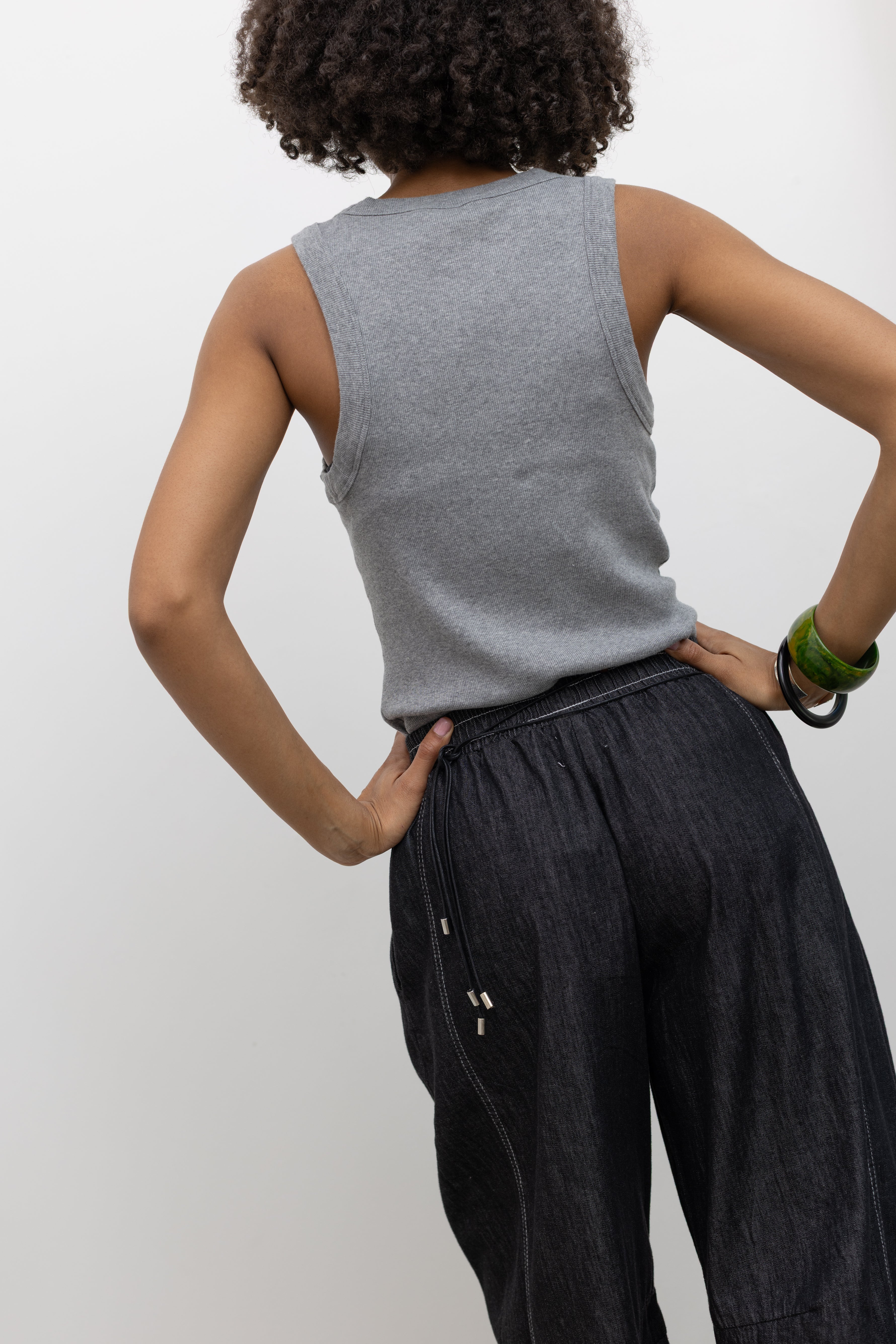 Model wearing an everyday essential in a Alas Eius label gray cotton blend ribbed tank top with contrasting embroidered AE logo on chest paired with Alas Eius Black Contrast Stitch Barrel Jeans and Veja slide sandals. 