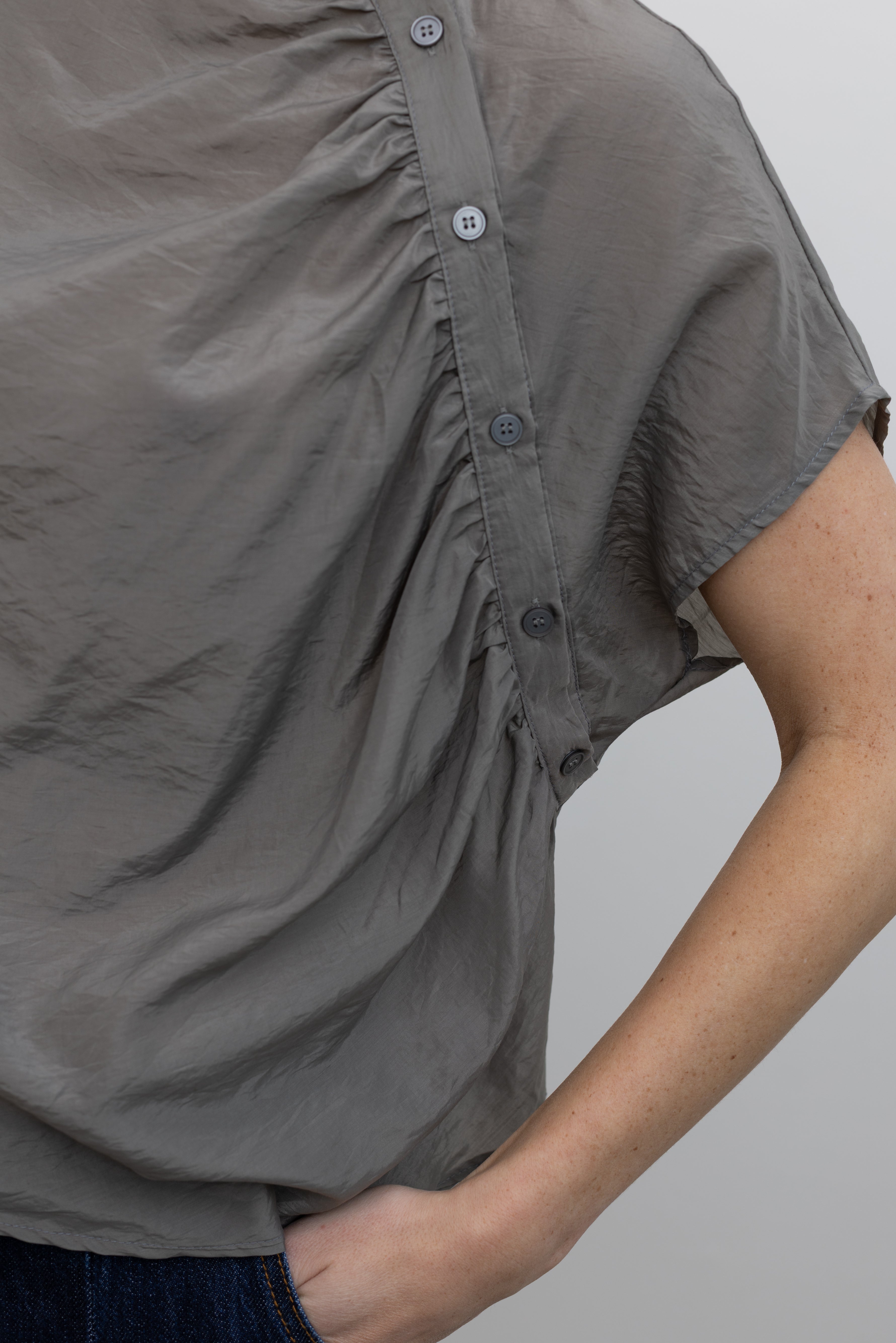 Close-up front view of a woman wearing modern minimalist spring fashion in an Alas Eius label light 
gray organza diagonal button front mock neck top with dark wash denim jeans at an Alas Eius store. 