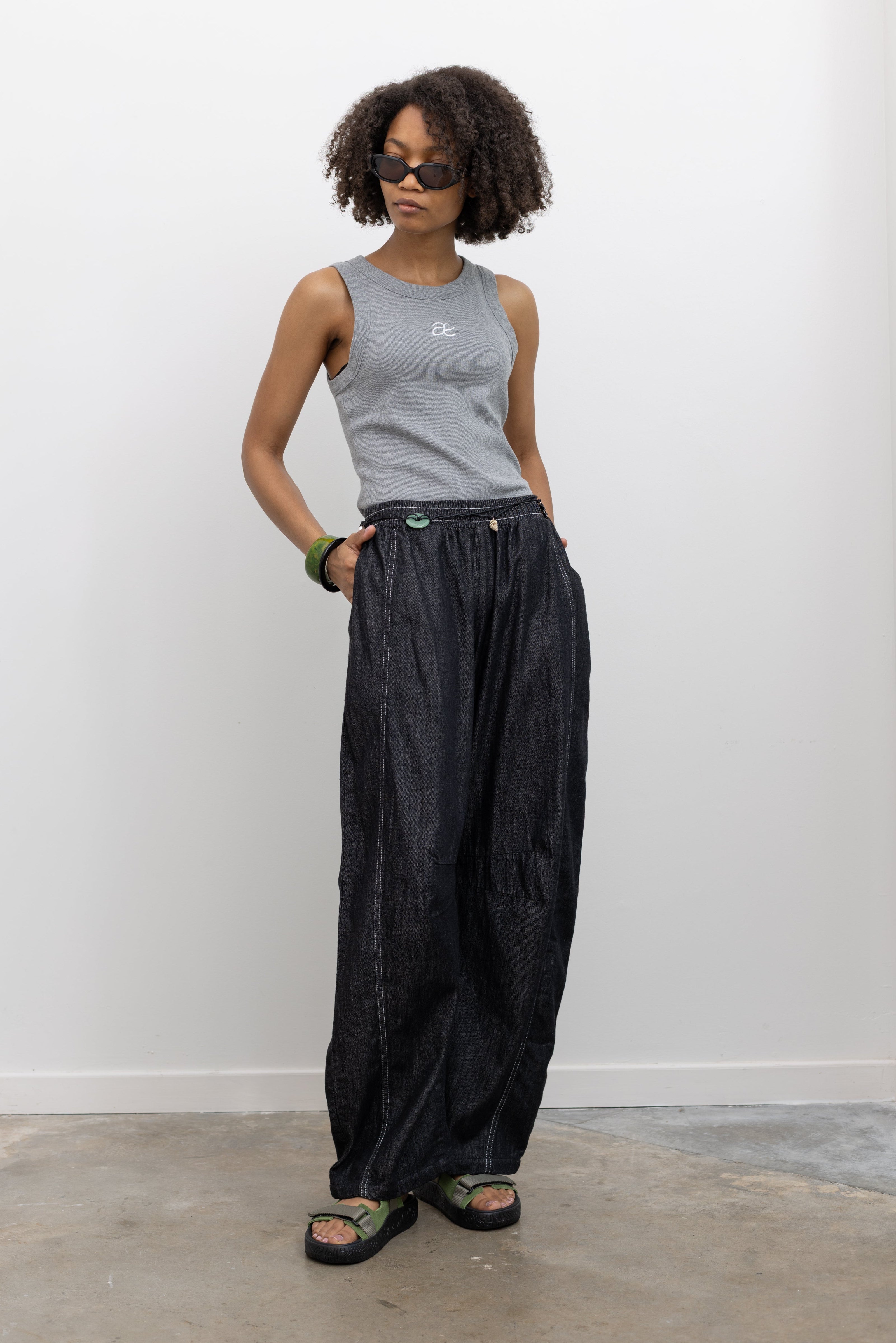 Model wearing an elevated basic in a Alas Eius label gray cotton blend ribbed racerback tank top with contrasting embroidered AE logo on chest paired with Alas Eius Black Contrast Stitch Barrel Jeans and Veja slide sandals. 