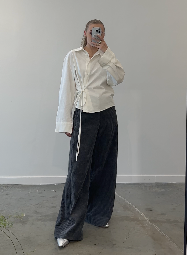 A person wearing an ivory button-down top with side ties and wide-leg black pants. The person is standing indoors, holding a mobile phone to their ear.