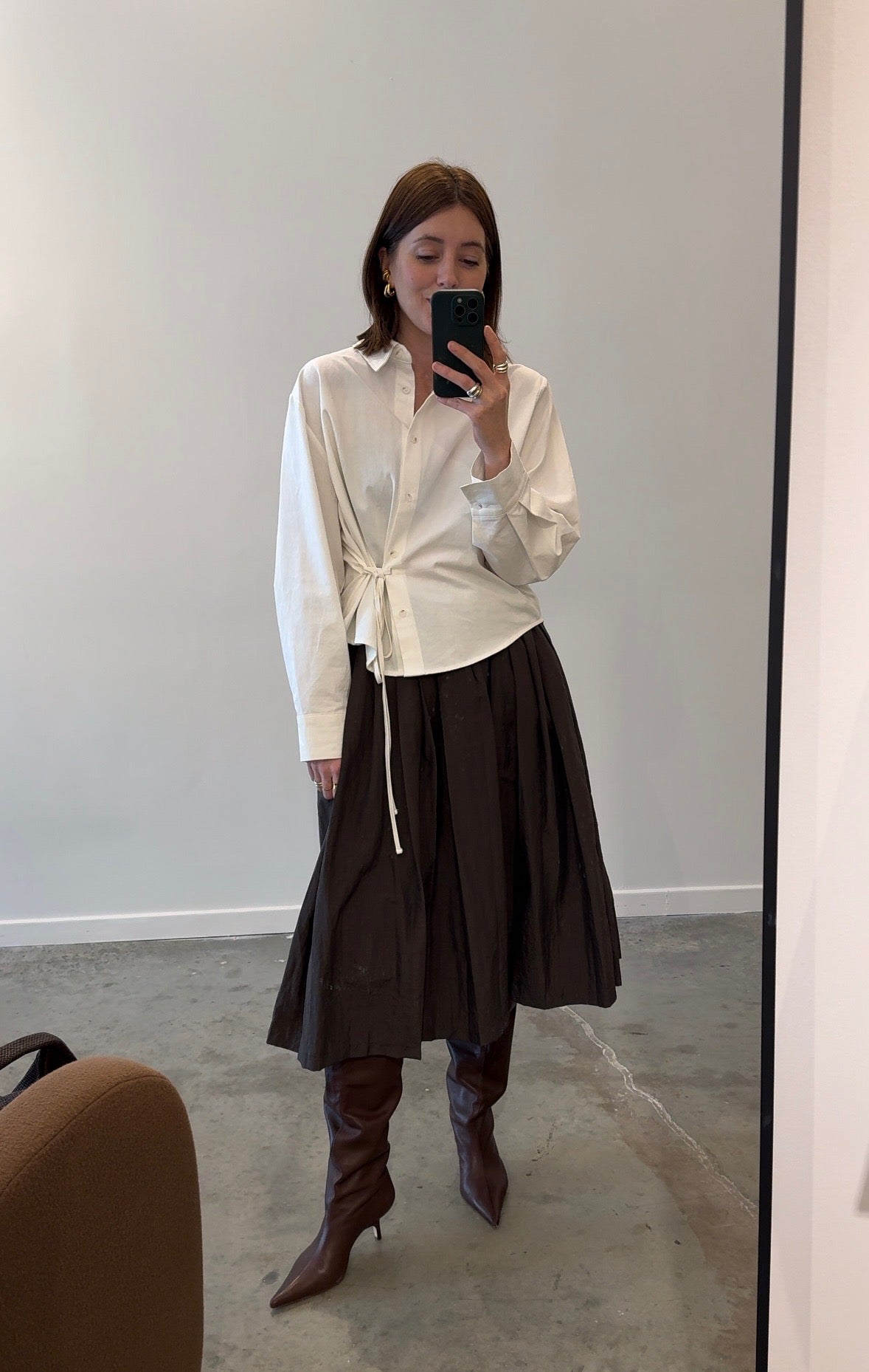 A person wearing a brown pleated full skirt with an elastic waistband, paired with a white top and brown boots. The skirt reaches mid-calf and is made of a lightweight fabric.