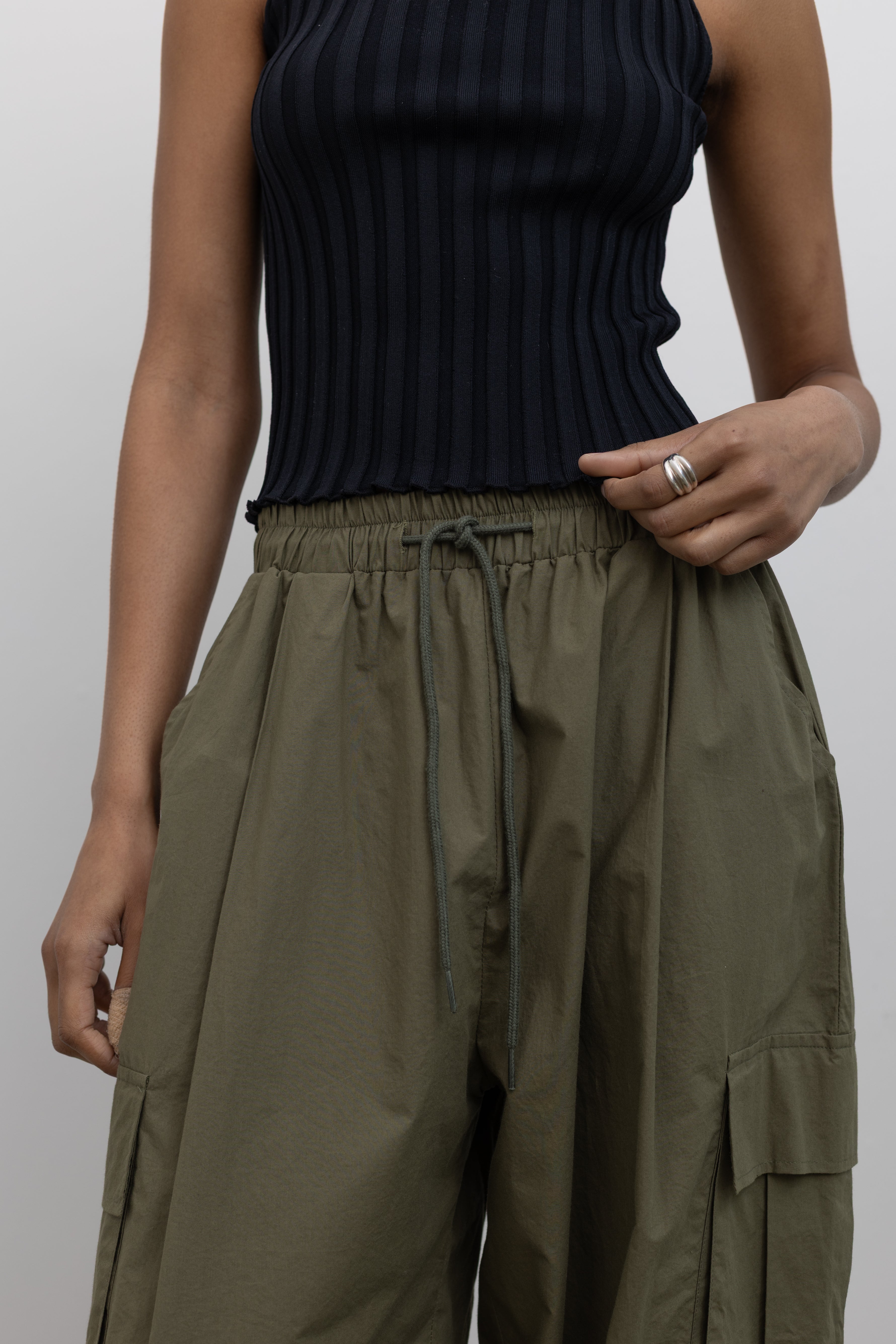 Product front view of Woman standing wearing everyday fashion in a pair of Alas Eius brand khaki green lightweight cotton wide-leg cargo pants with drawstring ankles and a tie waist and extra pockets at Alas Eius 