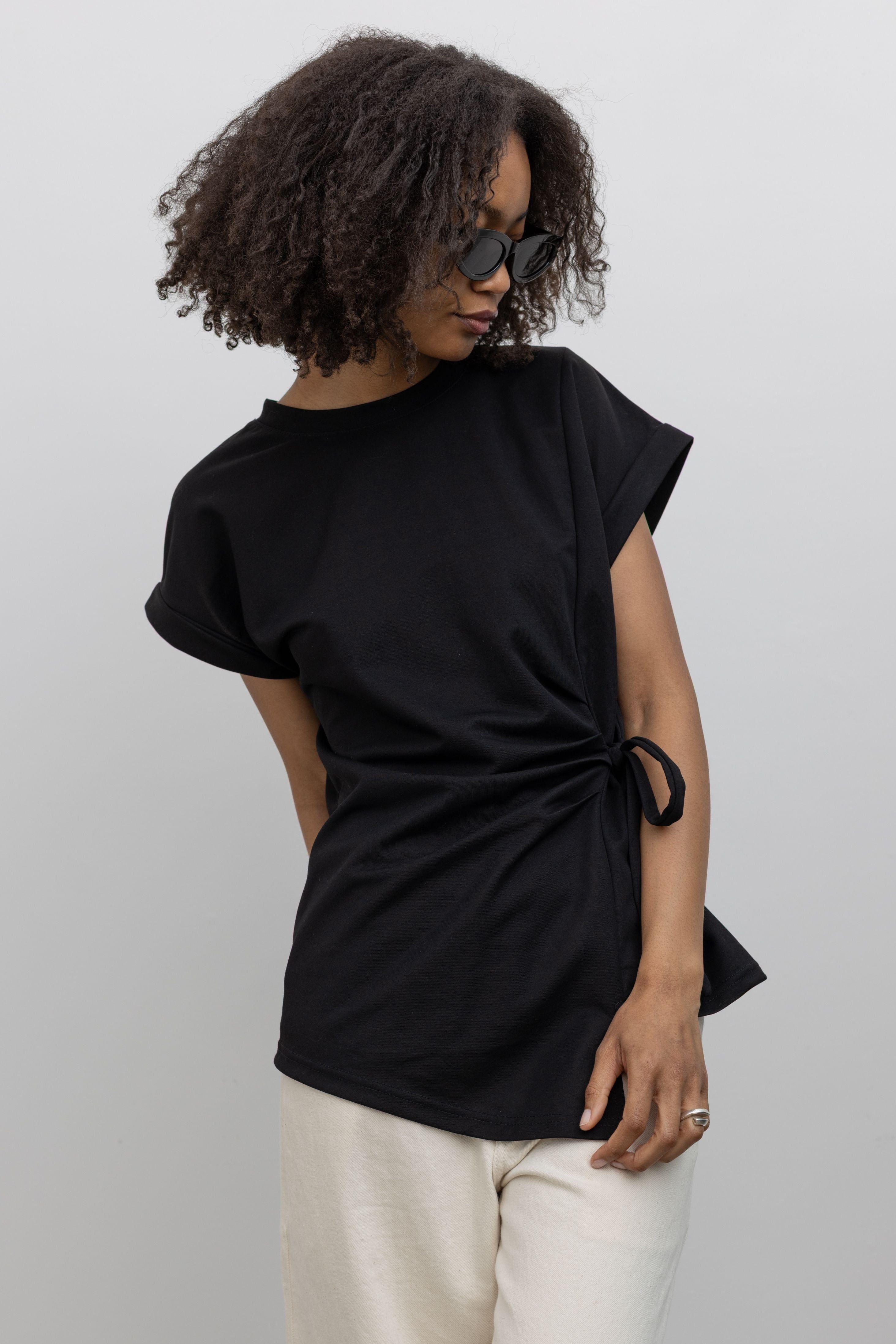 Front view of a woman wearing a wardrobe staple in the Alas Eius label black luxury cotton t-shirt with an asymmetrical side tie and rounded shoulders at an Alas Eius store. 
