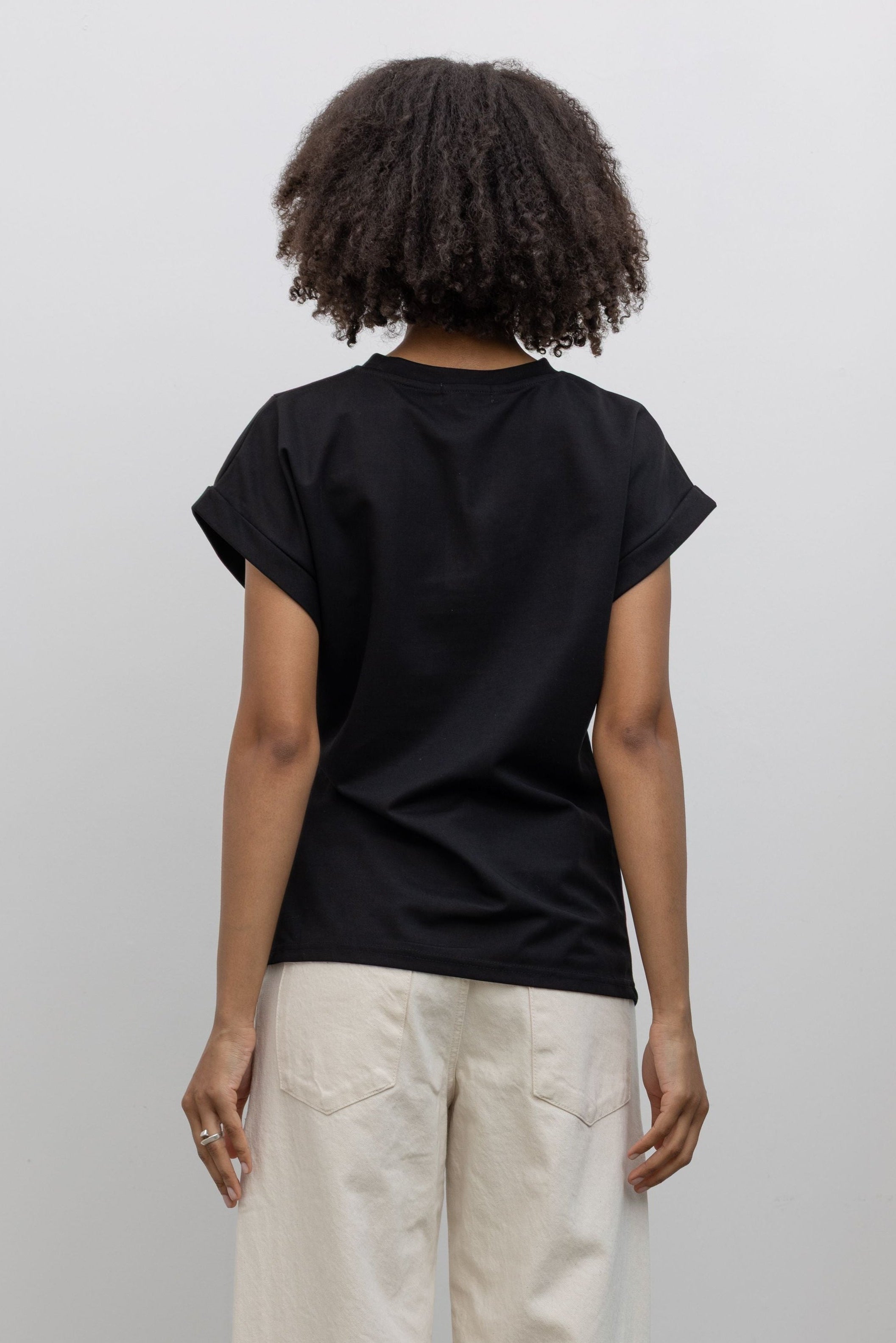 Back view of a woman wearing a wardrobe staple in the Alas Eius label black luxury cotton t-shirt with an asymmetrical side tie and rounded shoulders at an Alas Eius store. 