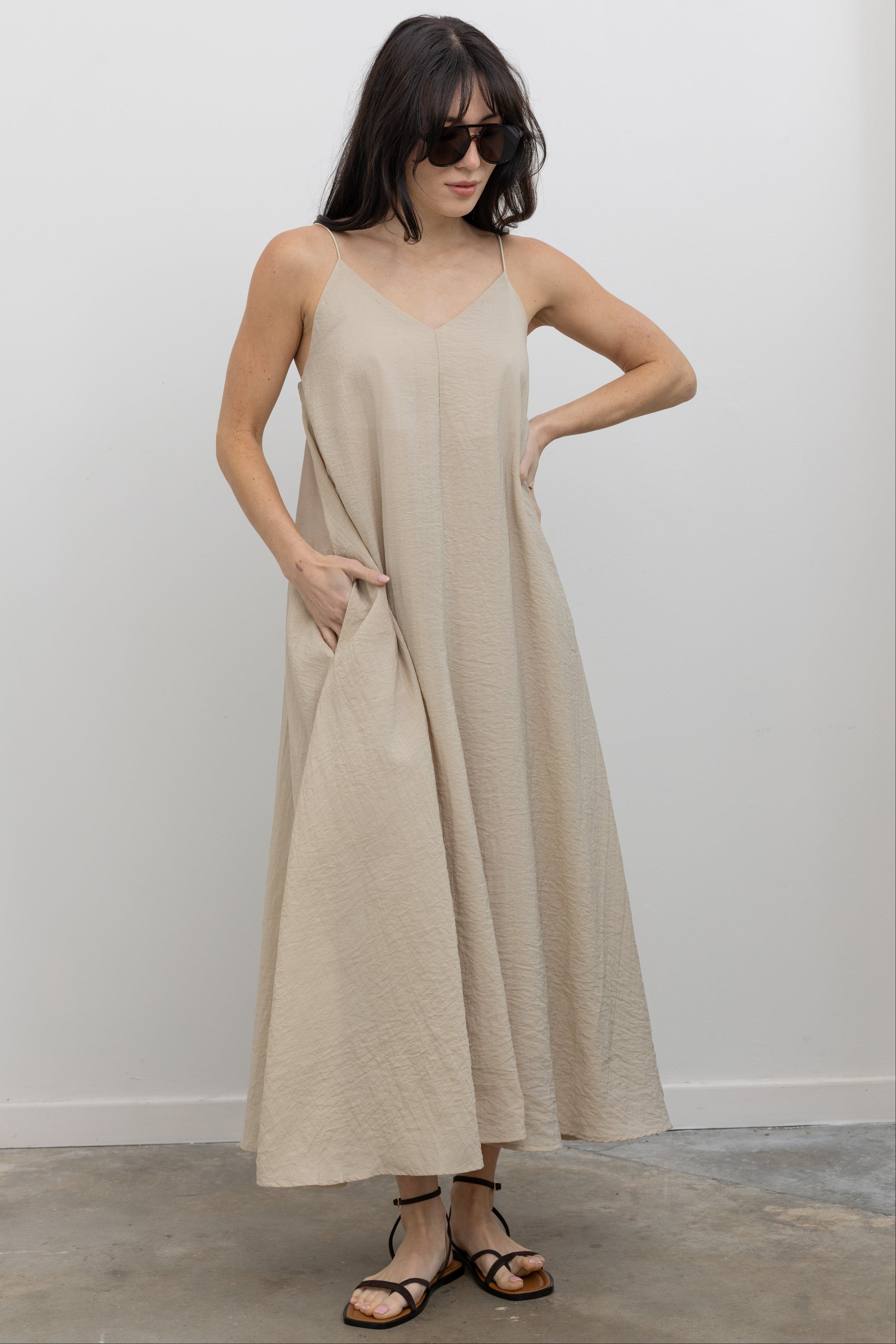 Full length front view of a woman wearing an elegant and flattering Alas Eius label spring and summer dress in a beige cotton blend featuring a flowing silhouette, adjustable bungee straps for a customized fit and two convenient side pockets paired with thin strappy black leather sandals at an Alas Eius store. 