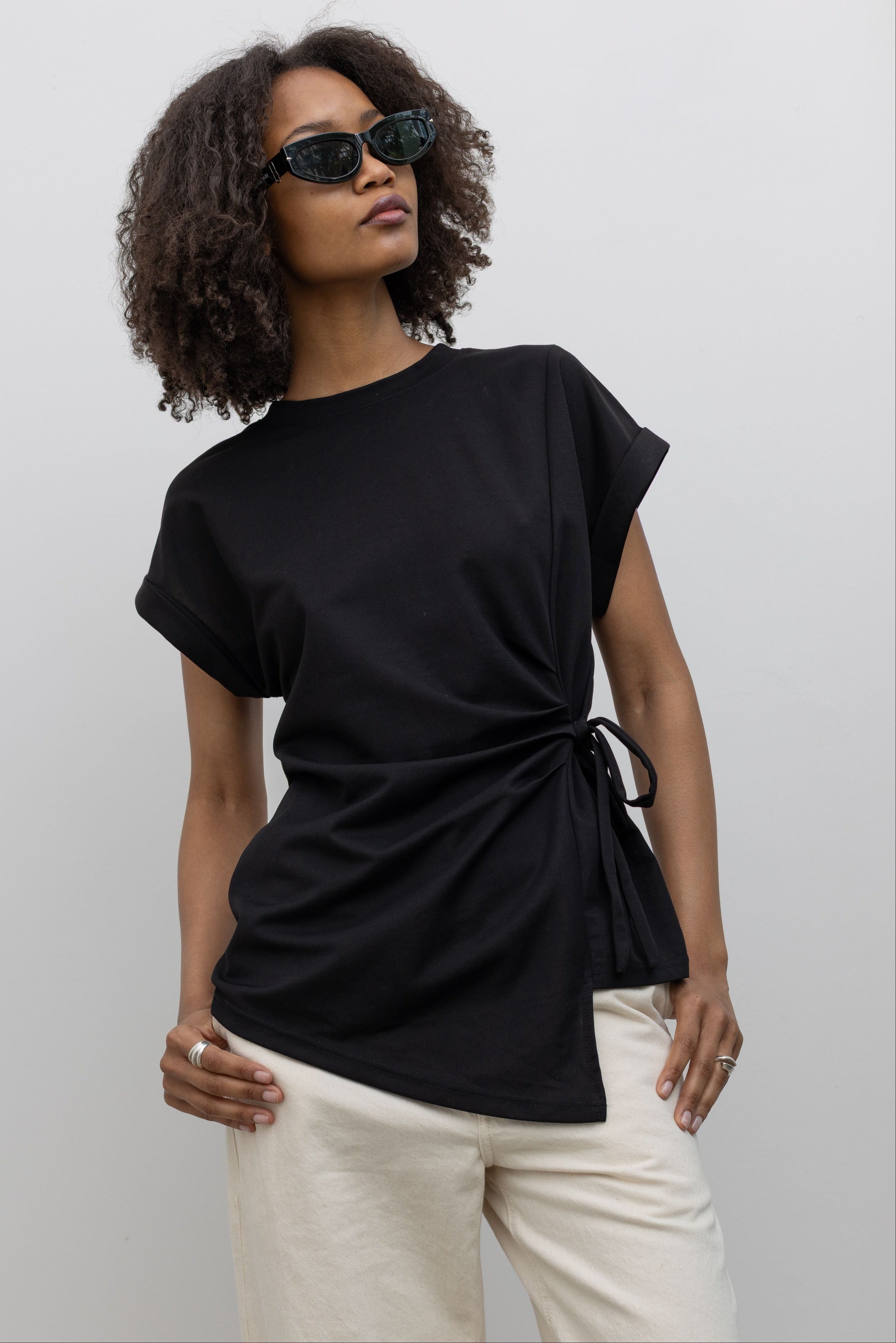 Front view of a woman standing wearing an elevated take on the black standard tee in an Alas Eius label black cotton blend side tie asymmetrical hem short sleeve top with white denim jeans at an Alas Eius store. 
